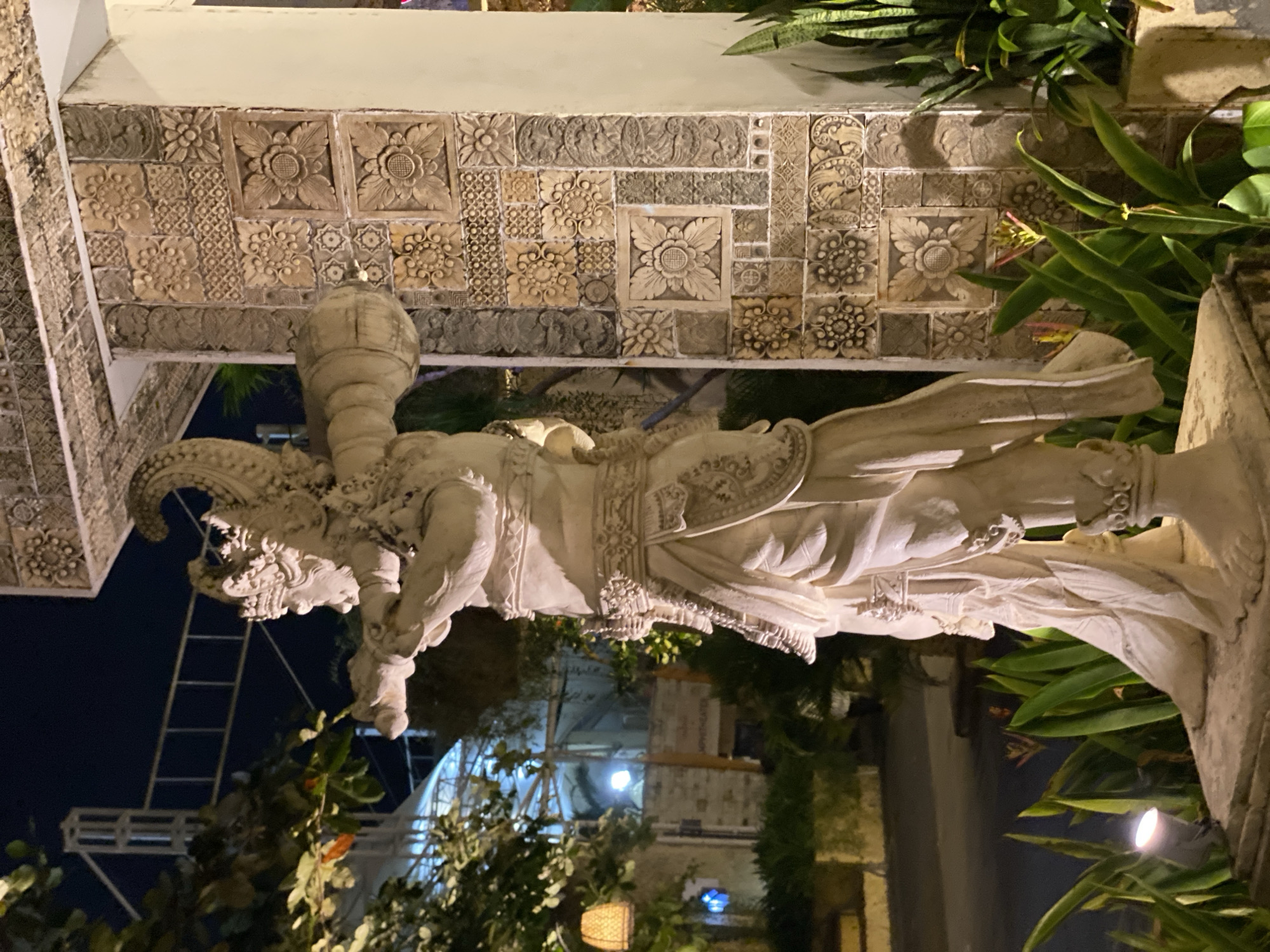 The media file shows a large stone statue of a mythological creature, likely a deity or guardian, in an outdoor setting at night. The statue is intricately carved, depicting a powerful figure with a fearsome face, elaborate headdress, and ornate clothing. It appears to be part of a decorative architectural element, possibly a temple or a grand entrance, given the carved stone panels on the wall behind it.
The background reveals a dark sky, indicating nighttime. Lush green foliage surrounds the statue, and artificial lighting illuminates the scene, casting shadows and highlighting the statue's details. In the distance, there are glimpses of building structures and what appears to be construction scaffolding, suggesting an urban or developing area. The overall atmosphere is somewhat mystical and imposing due to the statue's design and the nighttime setting. The location context provided, Kuta Selatan, Indonesia, points to a tropical environment where such ornate stone carvings are common in religious and cultural sites. No visible text is present in the image.
