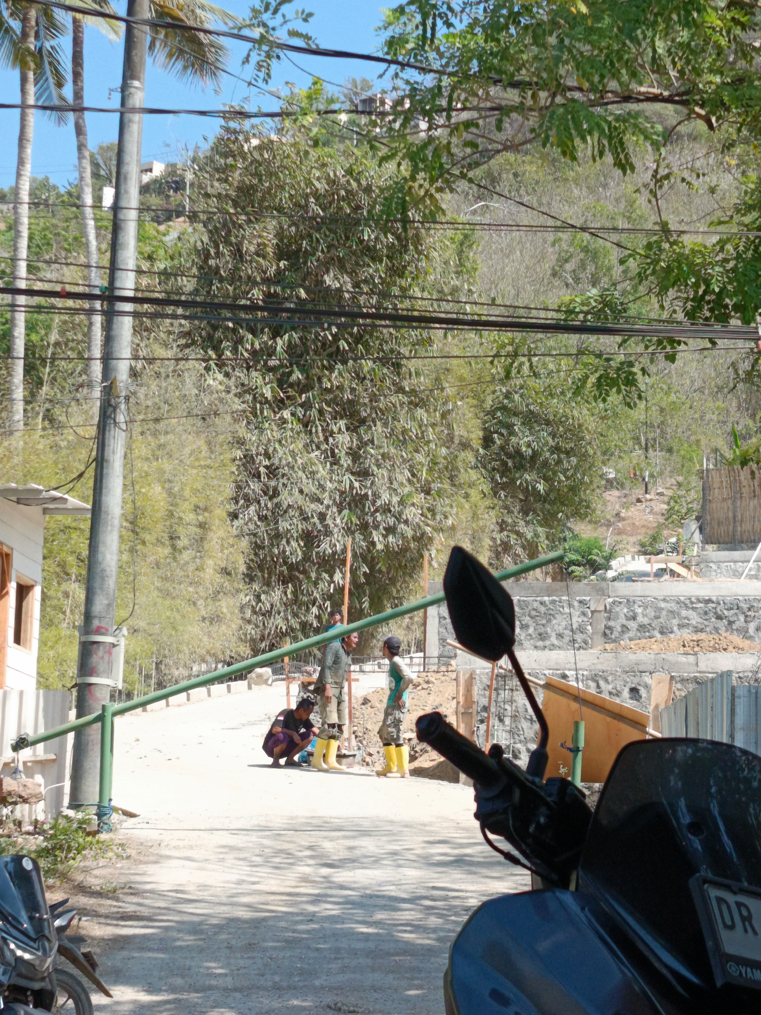 In Kuta, Indonesia, this image captures an outdoor scene with a dirt road leading to a construction area. A utility pole with overhead wires stands prominently on the left, adjacent to a white building with an orange-trimmed window. Several palm trees and dense bamboo foliage form the backdrop.
In the mid-ground, three individuals are present. One man, wearing a dark shirt and dark pants, is crouching. To his right, a man in a green long-sleeved shirt and light-colored shorts stands. Another person, partially visible, wears a green shirt and camouflage-patterned pants with yellow boots. A green metal gate obstructs the path.
The construction site features a retaining wall made of grey stones and concrete, with building materials and scaffolding visible. In the foreground, the handlebars and part of the seat of a dark-colored motorcycle are visible. The ground is uneven, with scattered rocks and a shadow cast across the road. The sky is clear and blue.