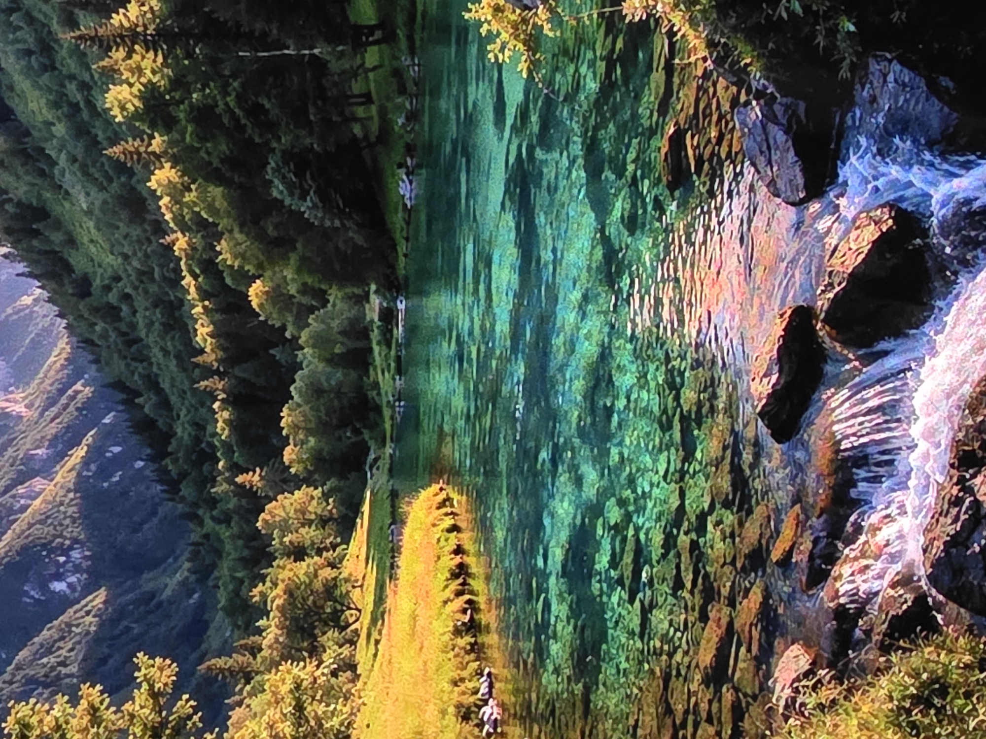 The image depicts a stunning natural landscape. A crystal-clear turquoise river flows from the foreground, revealing a rocky riverbed. Small rapids are visible in the lower part of the frame, with water cascading over smooth stones. Lush green trees line the banks of the river, extending up the steep slopes of a mountain range in the background. The mountains are covered in dense forest, with some areas showing rocky outcrops. Sunlight filters through the trees, illuminating patches of golden grass on the left bank and casting dappled light on the water. The overall scene conveys a sense of tranquility and pristine natural beauty. The location is listed as Unknown, Unknown.