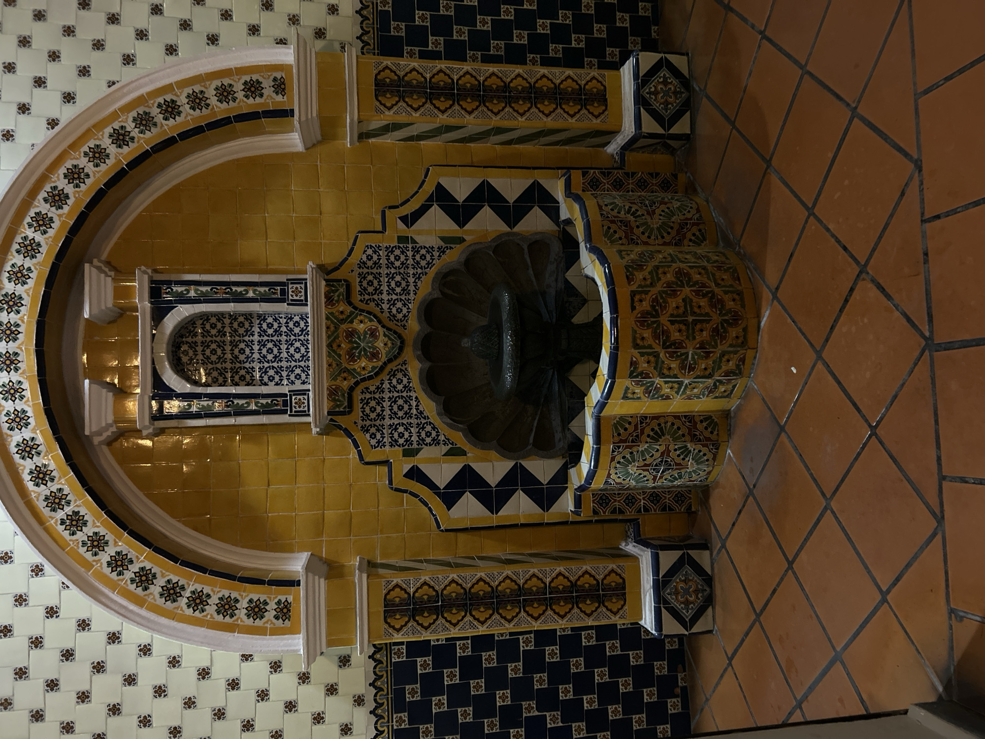 The image depicts a tiled water fountain in Santa Ana Yancuitlalpan (Chilhuacán), Mexico. The fountain is set against a wall covered in white and floral patterned tiles. A large, arched structure surrounds the fountain, decorated with yellow, white, and floral patterned tiles. The fountain itself is constructed with yellow and blue tiles, featuring a scalloped basin. The floor is covered with brown, diamond-patterned tiles.
