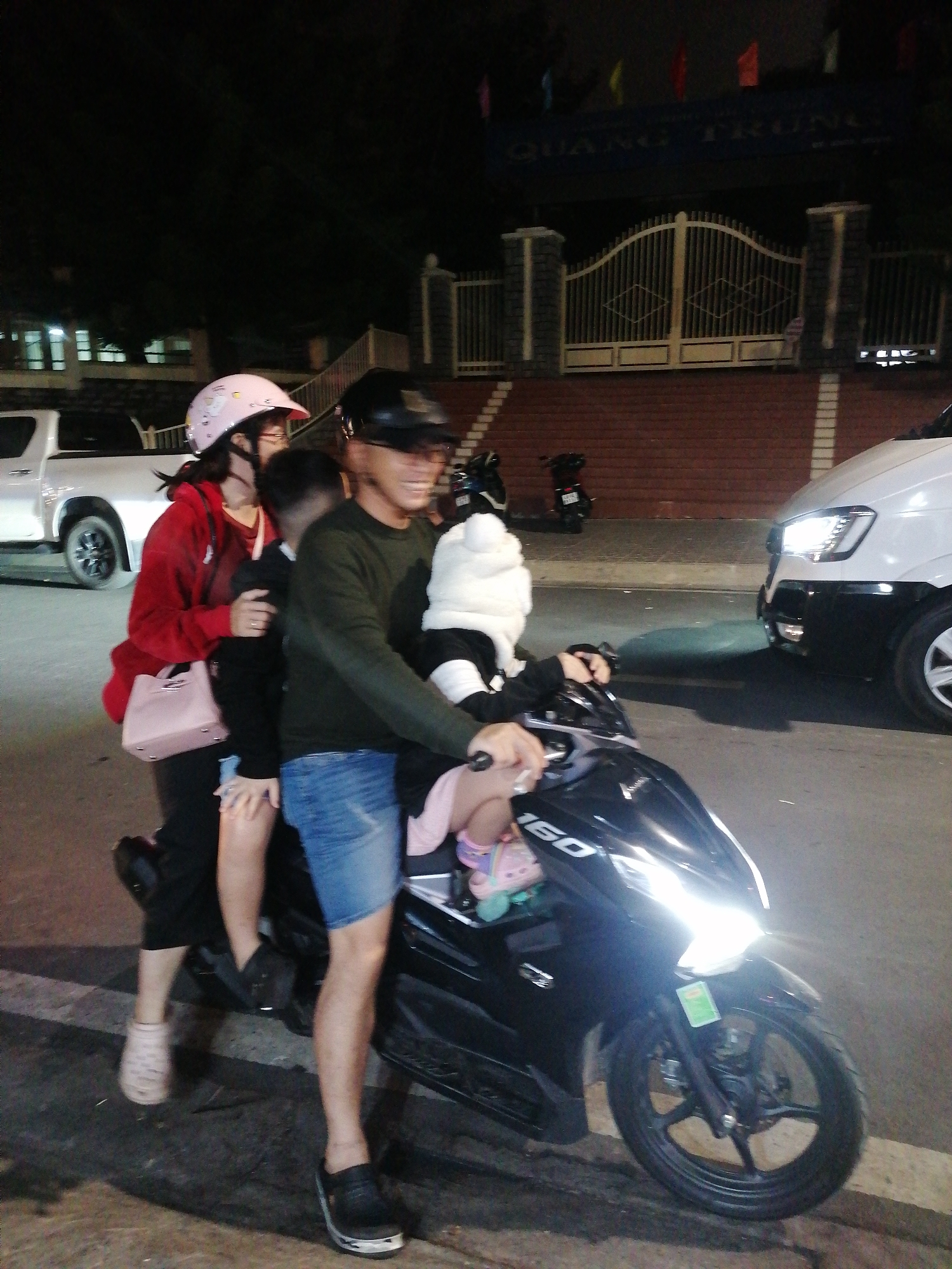 In Da Lat, Vietnam, on a paved road at night, three individuals are riding a black motorcycle. The person in the front is wearing a dark green long-sleeved shirt and denim shorts, and appears to be smiling. The person in the middle is wearing a red hooded jacket and a pink helmet adorned with a cartoon character. They are holding a light pink purse. A child is seated between the two adults. The motorcycle's headlight is illuminated, casting a bright light on the road. In the background, a white pickup truck is parked, and further beyond, a building with a large gate and decorative pillars is visible. The scene suggests a family outing or transportation at night.