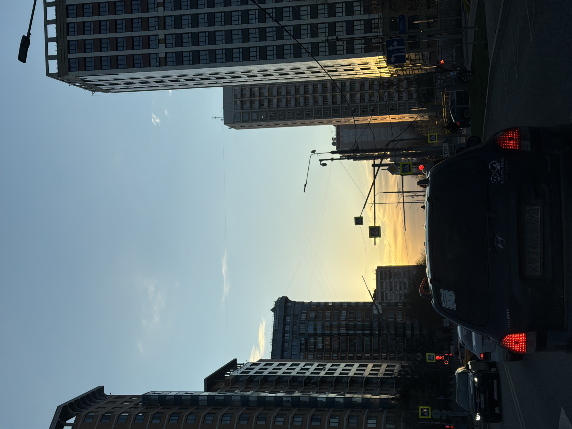 The image depicts a street scene in Moscow, Russia at dusk or dawn. The view is from within a vehicle, looking forward at a row of buildings on either side of the road. The buildings are multi-story residential complexes with repetitive window patterns. A dark-colored Hyundai vehicle is prominently visible in the foreground with its taillights illuminated. Several other cars are visible, also with illuminated lights, indicating that they are either stopped or moving slowly. Streetlights and traffic signals are present along the road. The sky in the background is a gradient of light blue and yellow, suggesting either a sunrise or sunset. The scene appears to be taken in a dense urban area, marked by residential buildings and traffic.