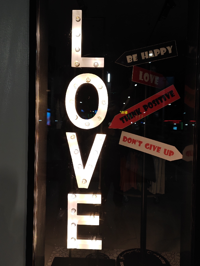 The scene displays a commercial storefront exhibit in Shumen, Bulgaria. A prominent vertical sign on the left spells "LOVE" using large, white, marquee-style letters, each letter internally illuminated by multiple visible light bulbs. To the right, four arrow-shaped placards are arranged diagonally downwards and to the right. From top to bottom, these placards read: "BE HAPPY" (black text on light ground), "LOVE" (white text on red), "THINK POSITIVE" (white text on red), and "DON'T GIVE UP" (red text on white). The entire display is viewed through a dark, reflective surface, likely a shop window, which shows indistinct reflections of ambient lights and internal merchandise, including clothing items on racks in the background. A partial dark, circular object bearing white text fragments, specifically "rega-tiv" and "@USE", is visible at the bottom right. No individuals are present.