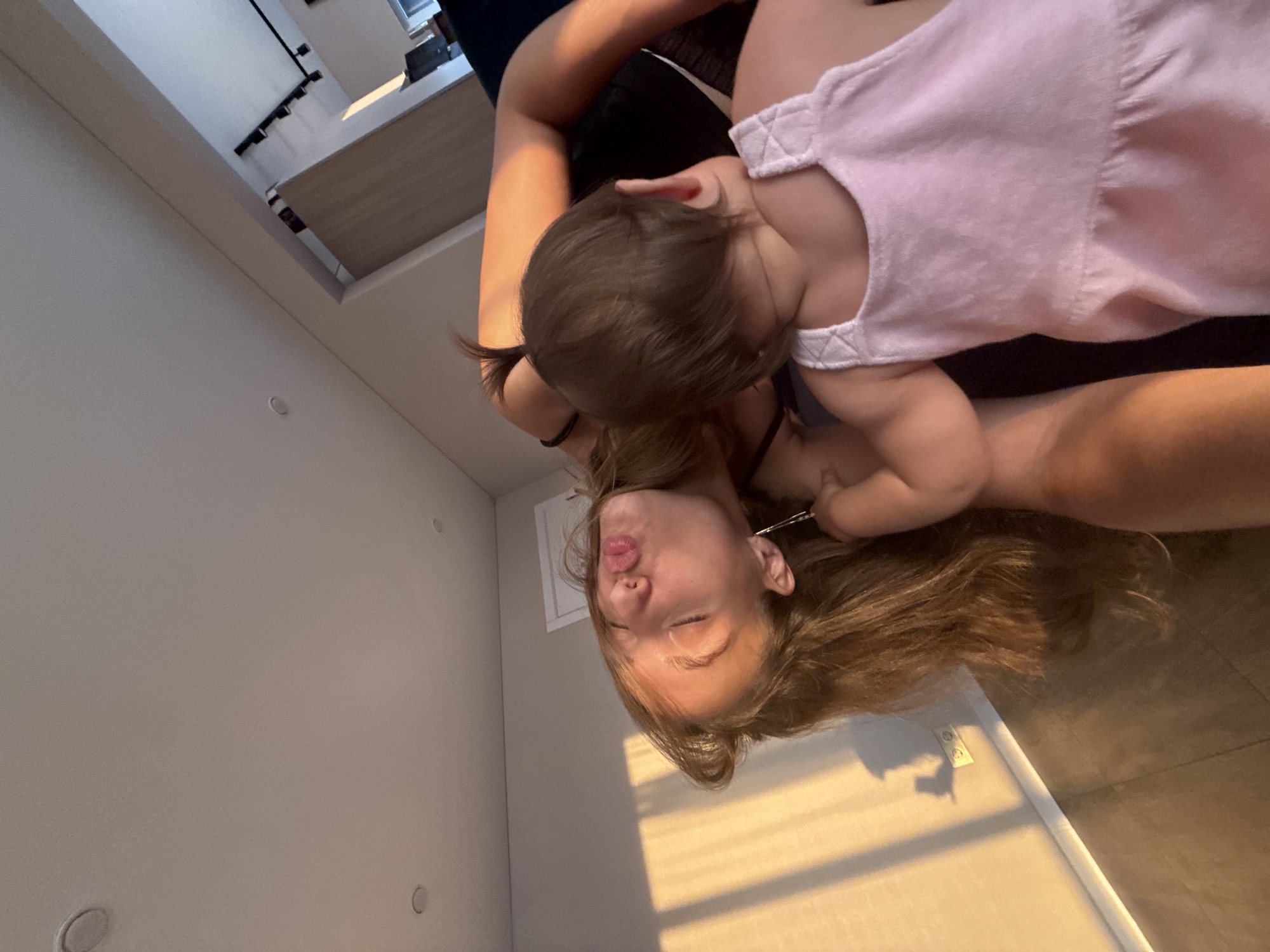 This media file shows a close-up, overhead shot of a person holding a baby. The person, who appears to be a young woman with long brown hair, is positioned below the baby. She is making a kissing face towards the camera, with her eyes closed and lips pursed. The baby is wearing a pink dress and is reaching towards the woman's face. The lighting suggests it is either early morning or late afternoon, with warm sunlight casting shadows on the wall behind them. The scene appears to be indoors, possibly in a home, with a plain wall and ceiling visible. The overall mood is playful and affectionate. No text is visible in the image.