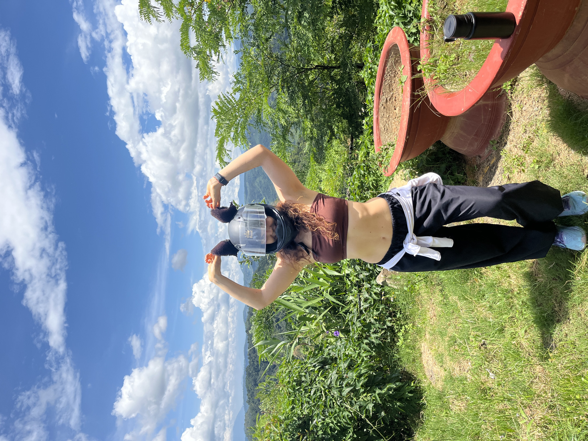 A woman with curly brown hair stands in front of a lush, green hillside with a blue sky and scattered clouds above. She is wearing a brown tube top, black pants tied with a white shirt, and light blue sneakers. A black motorcycle helmet with brown fuzzy ear attachments is on her head. She is holding her hands up, forming heart shapes above the helmet. Two large, terracotta planters are visible in the foreground, one with dry soil and the other with sparse green growth. A black water bottle rests on the edge of the planter with greenery. The woman is facing forward and smiling, with mountains and trees visible in the background. The scene appears to be a scenic overlook in Sơn Điền, Vietnam.