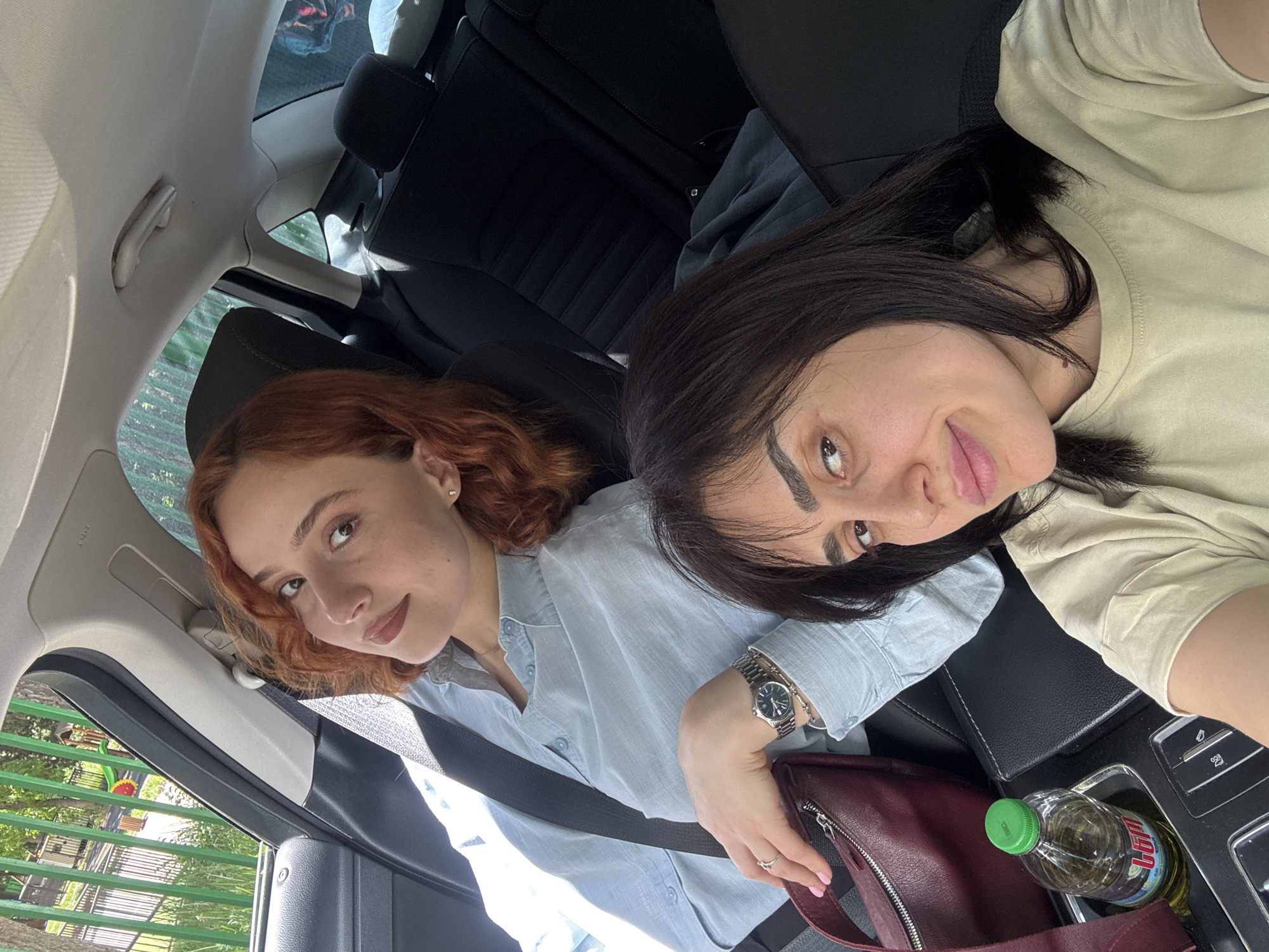Two women are sitting in the front seats of a car. The woman on the left has reddish-brown curly hair and is wearing a light blue button-down shirt with a seatbelt across her chest. The woman on the right has dark hair and is wearing a light tan t-shirt. She also has a seatbelt on, and a watch is visible on her left wrist. There's a maroon purse and a bottle of liquid in the car's console. Through the car window, a green park area is visible, with trees and a playground. Given the playground, park setting, and architectural styles, it's plausible the location is Tbilisi, Georgia.