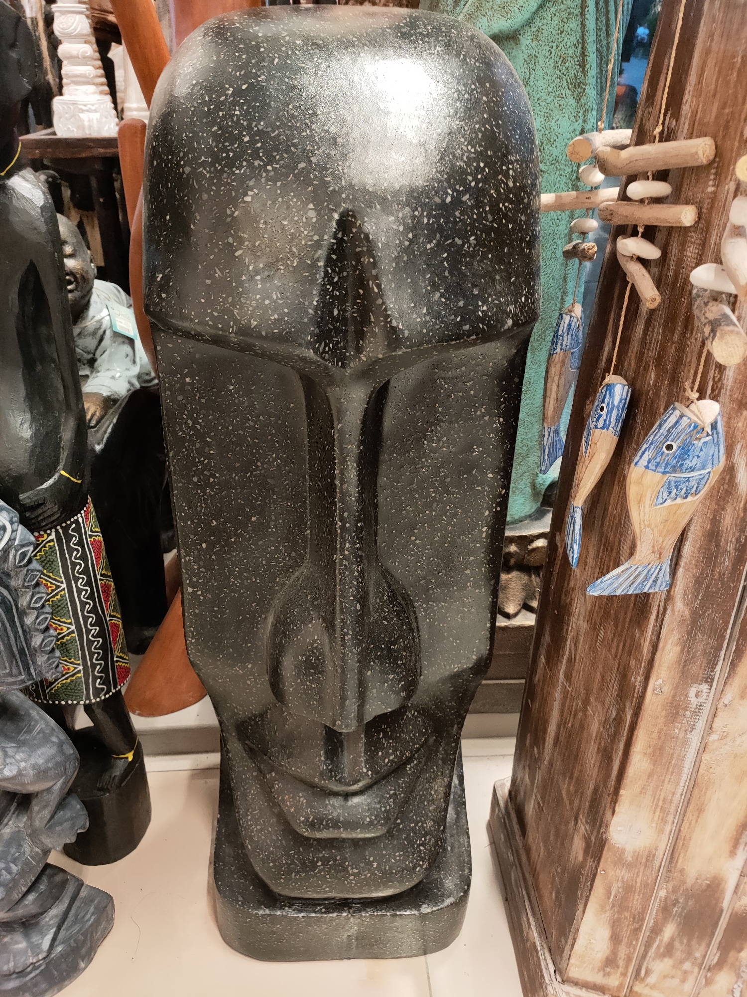 The image displays a close-up view of several decorative objects, likely in a shop or market setting. The main subject is a large, dark, stone-like sculpture of a stylized face with prominent features such as a long nose and a slight smile. The sculpture has a speckled texture, with small white and grey flecks embedded in the dark material. To the left of the main sculpture, a dark, carved wooden figure of a person is partially visible, adorned with a patterned fabric skirt. Further back, another smaller, dark figure, possibly a bust or statue, can be seen. To the right of the main sculpture, a wooden display stand is adorned with a mobile of small, carved wooden fish painted in blue and white, suspended by thin strings. The background shows other items, including what appears to be a light-colored ornamental column and a hint of a turquoise-colored surface. The lighting suggests an indoor environment. No text is visible in the image. The overall scene conveys a collection of artisanal or decorative items, potentially from a souvenir shop or craft market.