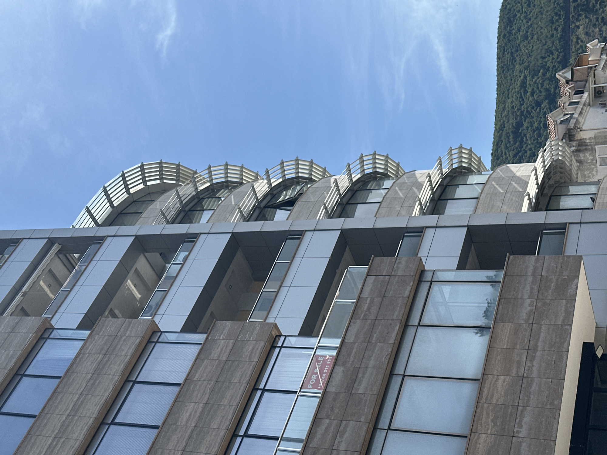 A low-angle shot features a modern architectural building under a clear blue sky with wispy clouds. The building is characterized by a stepped facade with alternating bands of stone and glass. A "FOR SALE" sign is visible in one of the glass windows. At the top of the building, a curved structure with a horizontal railing adds a distinctive design element. Part of another building with foliage is seen on the right side of the frame. This location is in the city of Budva, Montenegro.