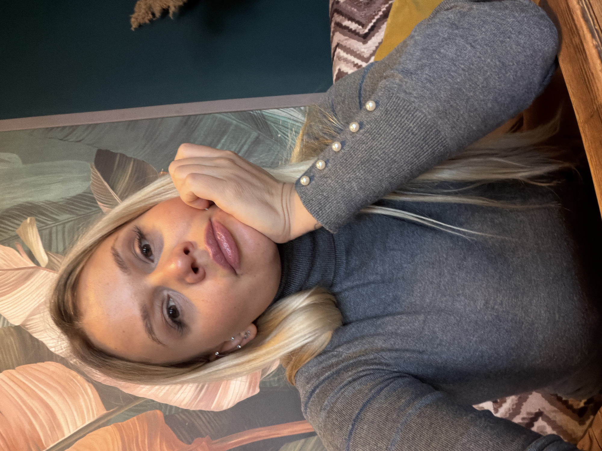 The image is a selfie-style photo of a blonde woman with fair skin. She is wearing a gray turtleneck sweater with pearl-like buttons on the sleeve. Her hair is light blonde with darker roots and is styled in a way that frames her face. She is resting her hand on her face. The background includes a wall with botanical-themed wallpaper in shades of pink, green, and brown, and a dark teal upper wall. A textured, dry floral decoration is also visible in the upper left corner. The setting is indoors, but the location in the city of Sarapul, Russia, cannot be determined from the image.