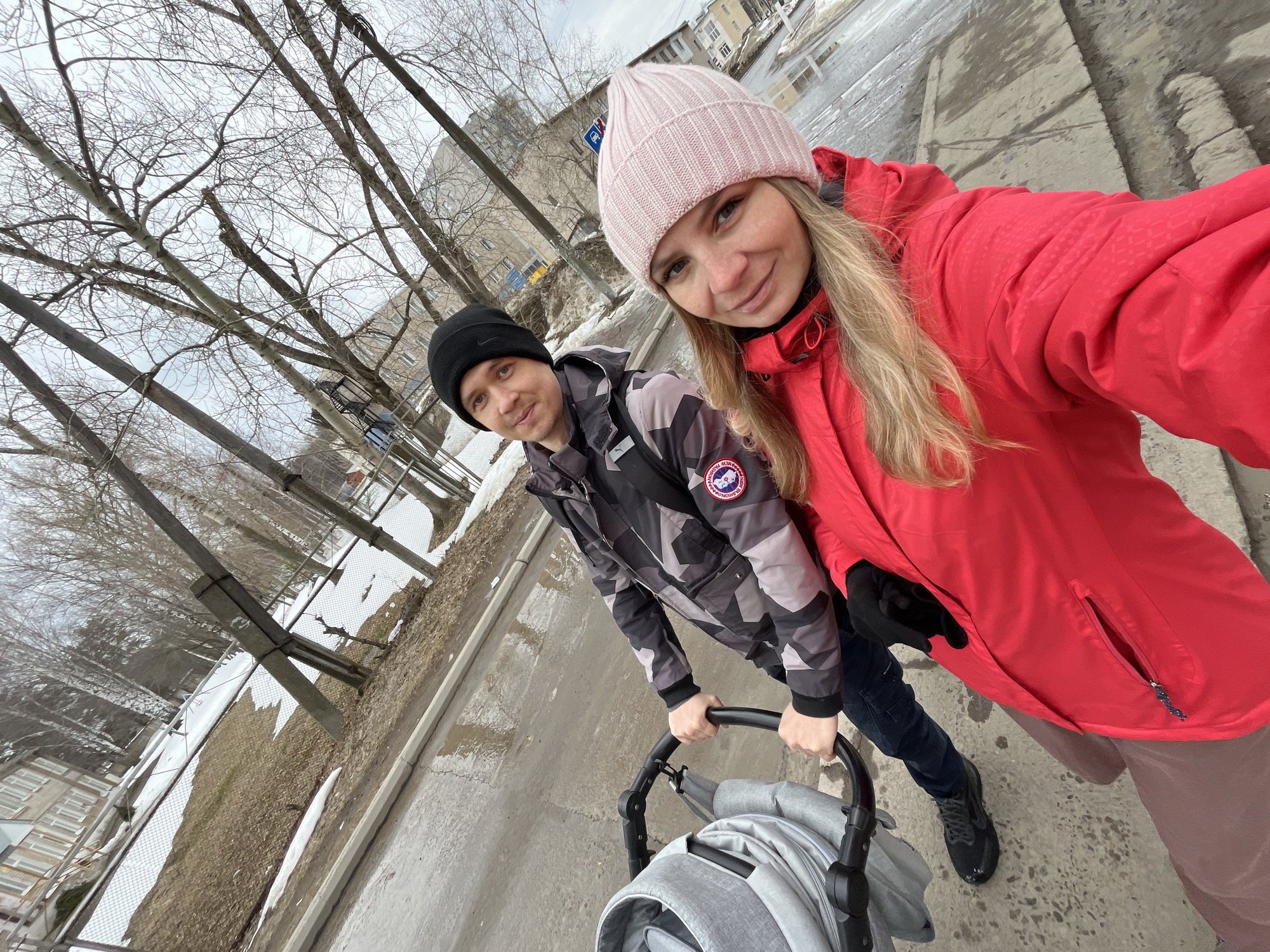 A couple with a stroller is on a city sidewalk in Chaikovsky, Russia. The woman wears a pink knit hat and a red jacket. The man wears a black knit hat and a gray camouflage jacket with a Canada Goose patch. They are pushing a gray stroller. The background shows bare trees, buildings, and patches of snow and ice on the ground and sidewalk, indicating early spring conditions. The road is wet. The photograph appears to be a selfie taken by the woman.