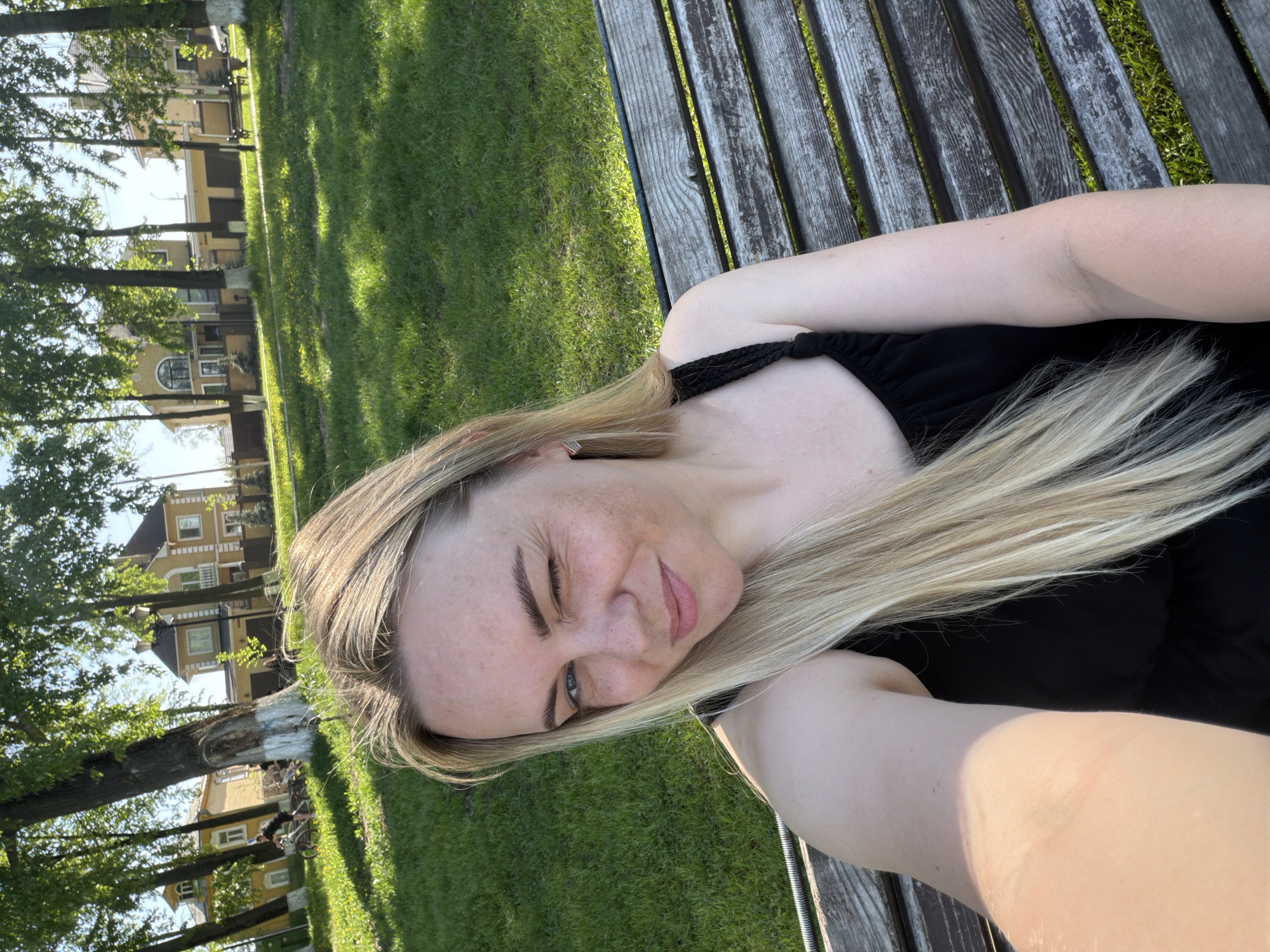 A woman with blond hair is lying on a wooden bench outdoors. She is wearing a black top. Behind her is a grassy area with trees and buildings in the background. The location is possibly a park or recreational area in a city. The style of the buildings and trees suggests a location in Russia.