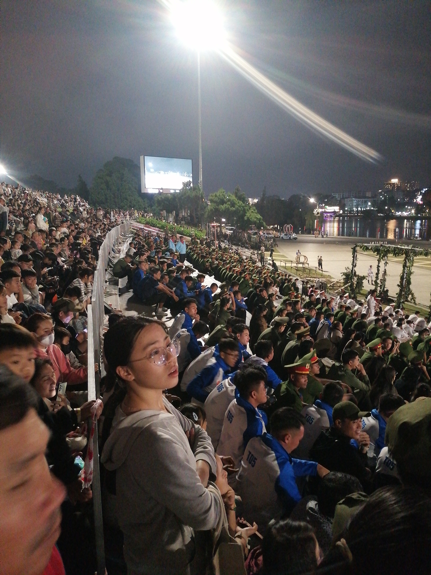 The image depicts a nighttime outdoor event in Da Lat, Vietnam, likely a performance or ceremony. A large crowd is seated in bleachers, facing a stage area that is not fully visible. The atmosphere is illuminated by bright spotlights and a large screen displays images. In the foreground, several individuals are visible, including a woman in glasses looking off to the right. The crowd appears to be a mix of civilians and individuals in uniform, possibly military or cadets, some wearing green caps and jackets. The background reveals a body of water with lights reflecting on its surface, suggesting proximity to a lake or river, common in Da Lat. Trees and buildings can also be seen in the distance, characteristic of the city's landscape. The overall scene conveys a sense of public gathering and organized entertainment.