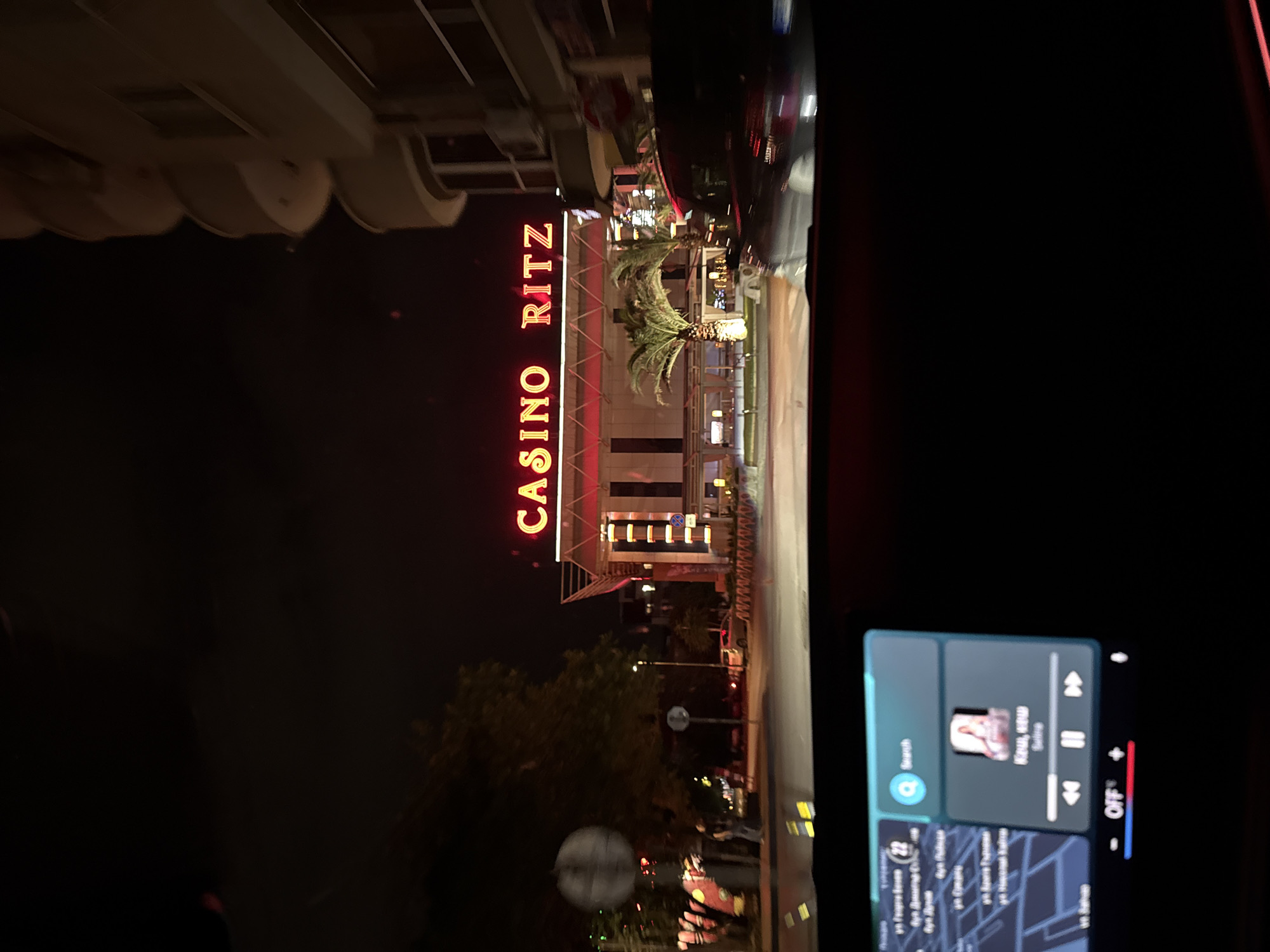 The image captures a nighttime street view from inside a vehicle, showcasing the illuminated facade of the "CASINO RITZ" building. The casino's name is prominently displayed in large, bright red neon letters, oriented vertically on the building's side. In front of the building, several palm trees are visible, lit by the surrounding lights. The scene is dark, typical of night, with various light sources emanating from the building and street.
In the foreground, to the lower right, a section of the car's infotainment screen is visible. This screen displays a music player interface, showing "KARA, KENU" as the artist and "Corine" as the song title. Below the music player, a map application is displayed, featuring street names written in Cyrillic script. Notable visible street names include "ул. Георги Сава Раковски" (ul. Georgi Sava Rakovski), "ул. Патриарх Евтимий" (ul. Patriarh Evtimiy), and "ул. Княз Александър I" (ul. Knyaz Aleksandar I), which are common in Bulgarian cities like Plovdiv. The presence of Cyrillic text and the specific street names confirm the location within Bulgaria. The activity depicted is being a passenger or driver in a car at night, passing by or parked near the casino.