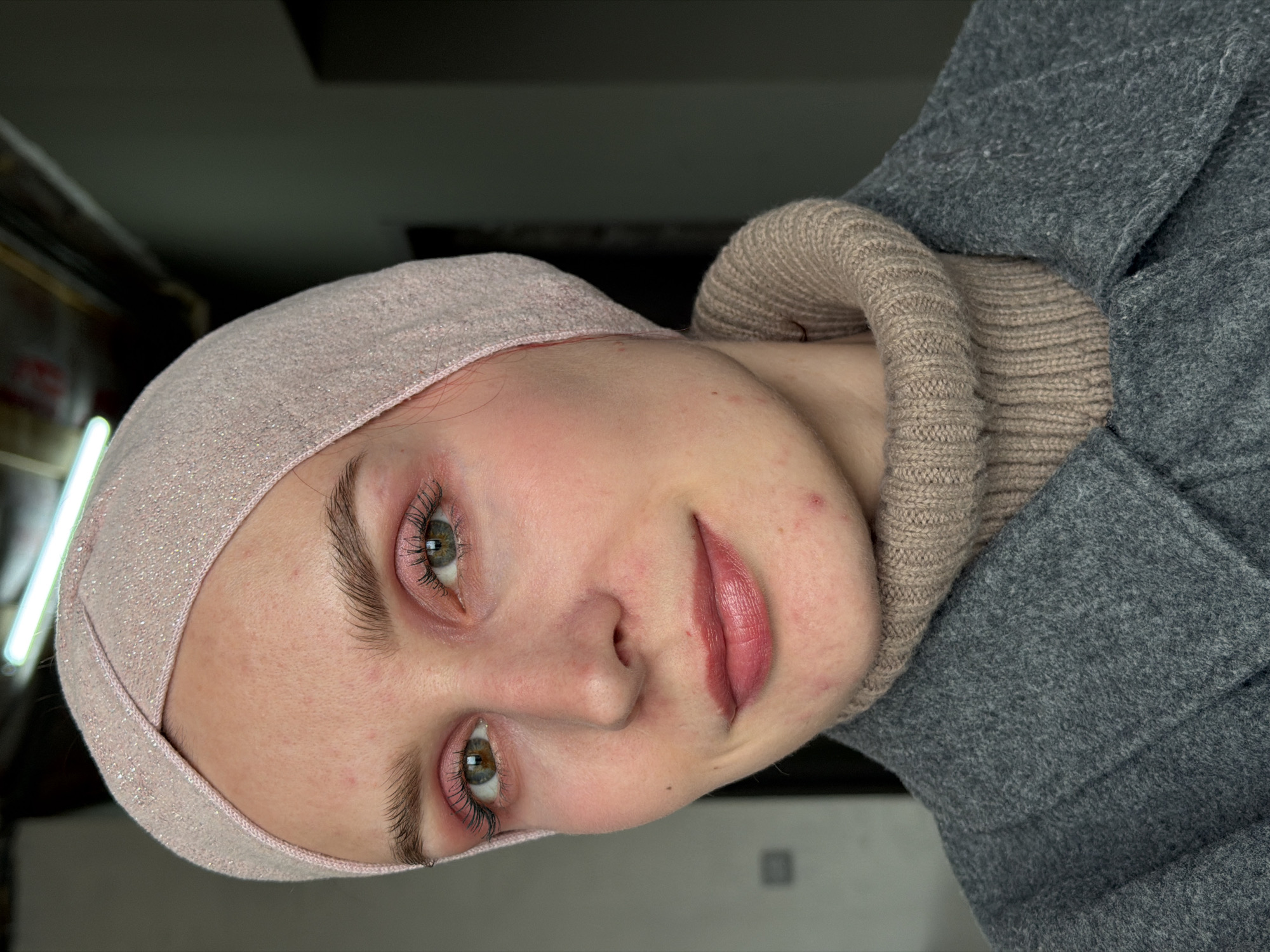 A young woman with fair skin, wearing a light pink head covering and a beige turtleneck sweater underneath a grey coat, is depicted in a close-up portrait. The photograph is taken indoors. Her eyes are green, and she is wearing light pink lip color. There is no indication of the location being Chaikovsky, Russia, visible in the image.
