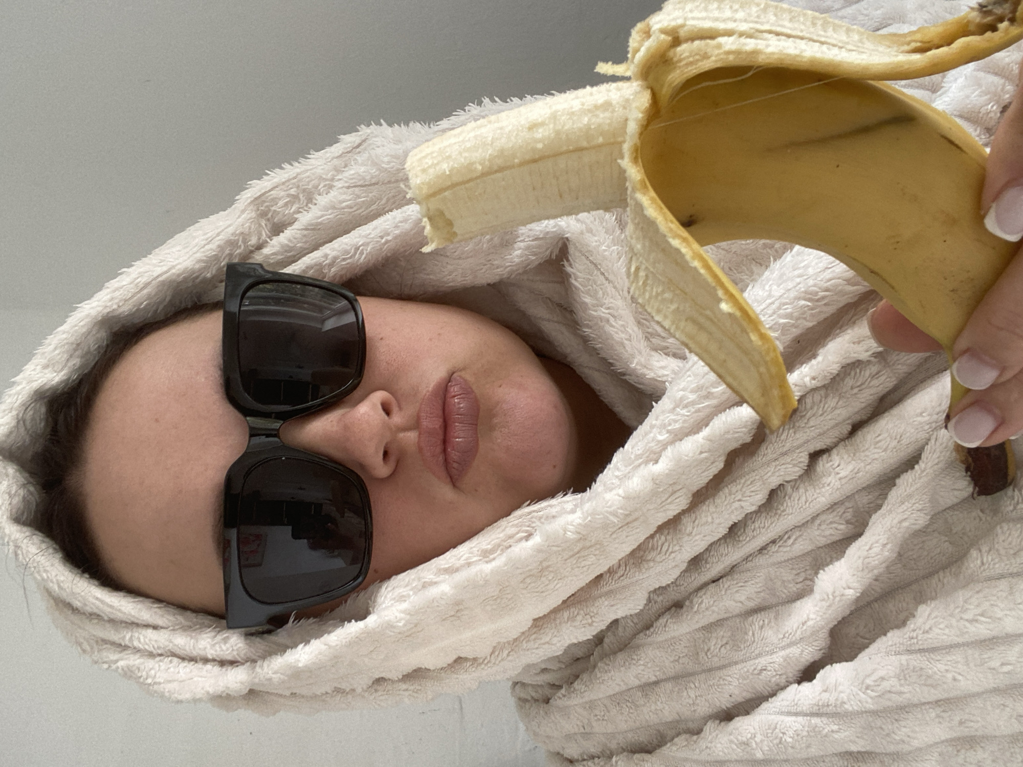 A person with sunglasses and a white towel wrapped around their head is holding a peeled banana. The person's face is visible, and their lips are prominent. The background is a plain white surface. The photograph appears to be taken indoors, but there is no indication of it being in Sochi, Russia.
