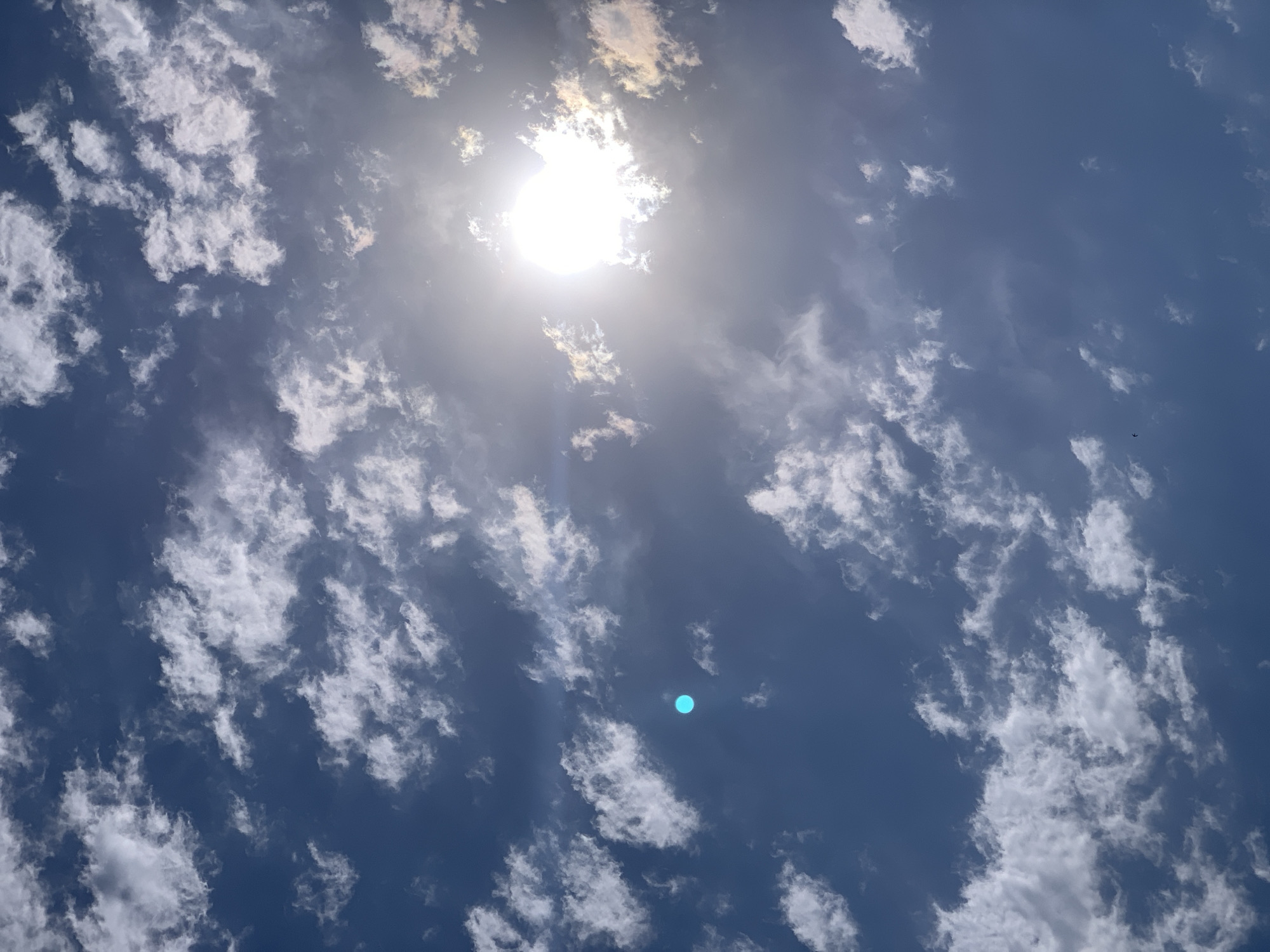 The image shows a bright sun partially obscured by wispy white clouds against a deep blue sky. A distinct, small, bright cyan circle is visible in the upper left quadrant of the sky. The sun is positioned in the upper right quadrant, emitting strong rays and lens flare. The scene appears to be an outdoor, aerial view, suggesting it was taken during daylight hours. There are no people, buildings, or other identifiable landmarks present in the image. The environment is solely composed of the sky, clouds, and the sun.