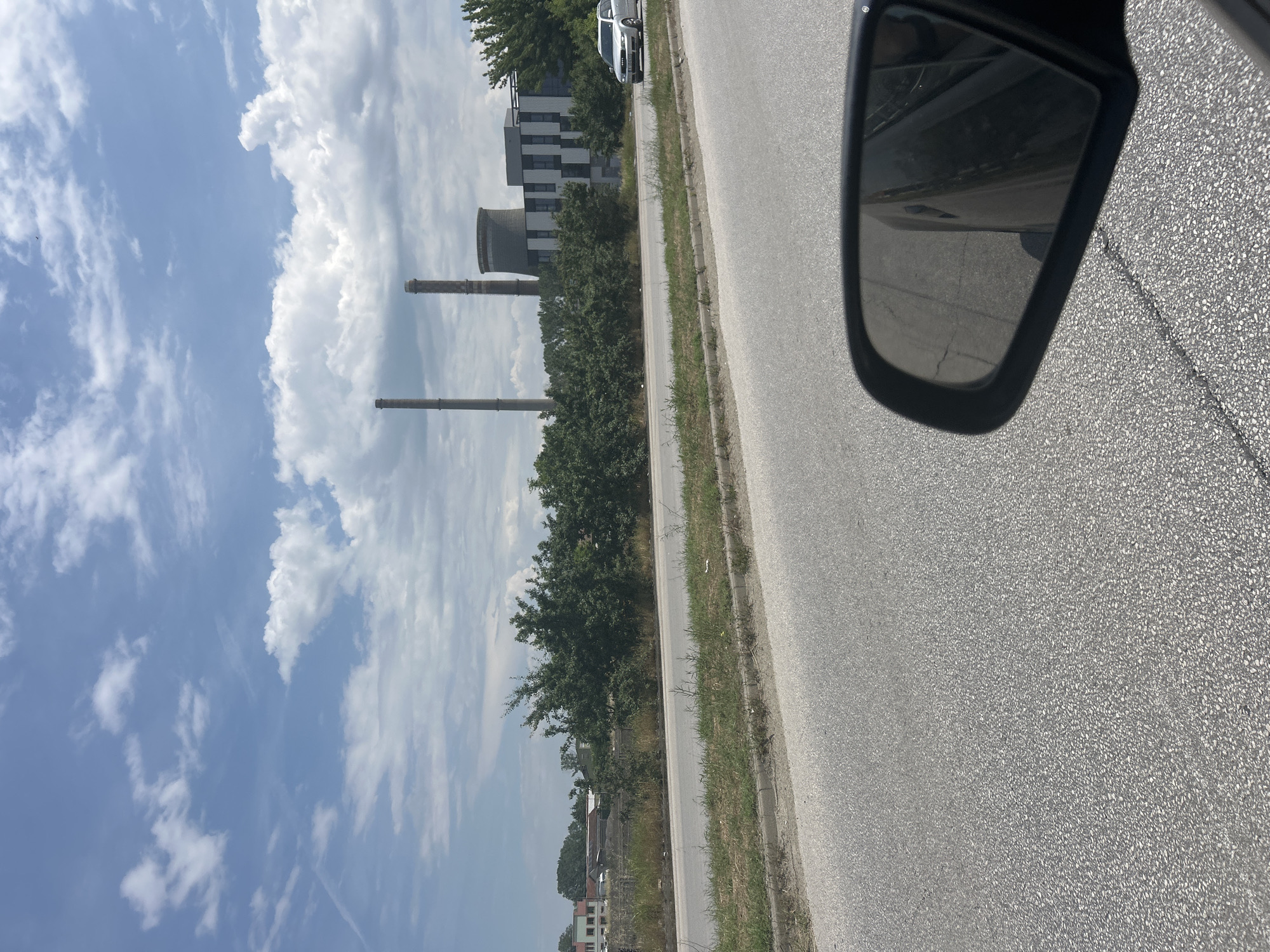 The image, taken from inside a vehicle, presents a bright daytime scene alongside a road. An asphalt road, bordered by a grass verge, occupies the right side of the frame, receding into the distance. A black car side mirror is visible in the upper right corner, reflecting a dark interior or side of the vehicle and a small portion of the road.
To the left of the road, a line of green trees and bushes partially screens the background. Beyond these, the scene features prominent industrial elements: two tall, slender grey chimneys or smokestacks, and a large, hyperboloid-shaped cooling tower, suggesting a power plant or significant industrial facility. Adjacent to these structures, a modern multi-story building with a distinctive black and white patterned facade stands. A silver car is parked on the side of the road further down near this building.
The upper and left parts of the image show a bright blue sky dotted with scattered white cumulus clouds, indicating fair weather. The overall setting appears to be an industrial or semi-urban area, observed during travel. No visible text is present. This scene is located near Plovdiv, Bulgaria.