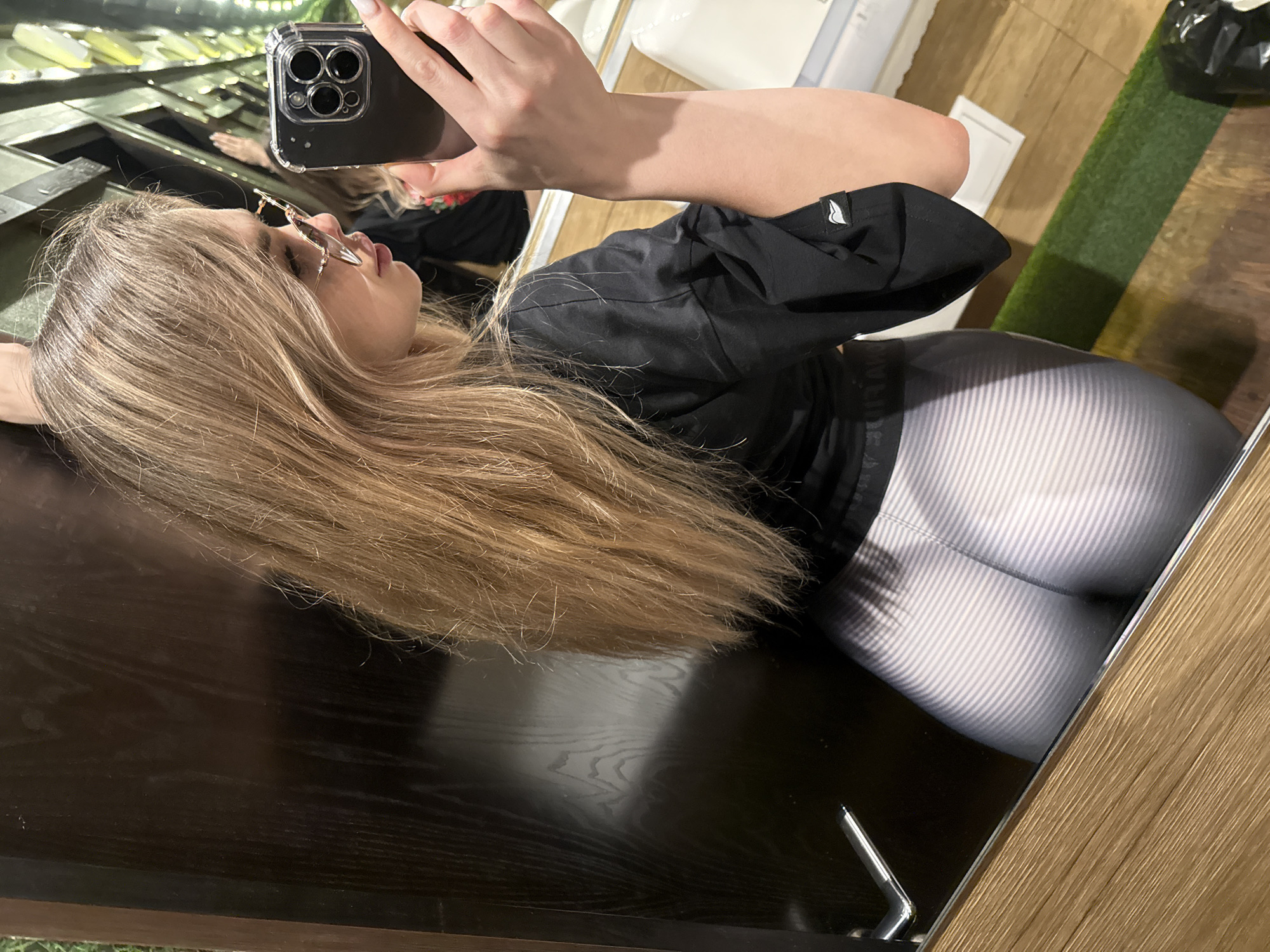 An image captures a mirror selfie of a woman lying down indoors. She is positioned on her side with her head and long, light brown hair resting on a dark, highly reflective surface. Her right arm is extended, holding a smartphone with multiple cameras and a clear case, aiming it towards her reflection to take the photo.
The woman wears distinctive, possibly cat-eye or butterfly-shaped sunglasses. Her face is seen in profile, with her mouth slightly open. She is dressed in a black top or jacket that features a small, white, stylized logo on its left sleeve, resembling a bird or a wave. Her lower body is covered by form-fitting leggings with a prominent pattern of diagonal white and grey stripes.
The background, visible through the reflections, reveals a diverse indoor setting. Elements include sections of light wooden flooring or paneling, a strip of green artificial turf, and what appears to be light-colored furniture or objects. The dark surface she is resting on provides a clear reflection of herself and parts of the room. The lighting suggests an indoor environment, but the specific time of day is not discernible. No explicit textual information is clearly visible, apart from the stylized logo on her sleeve.