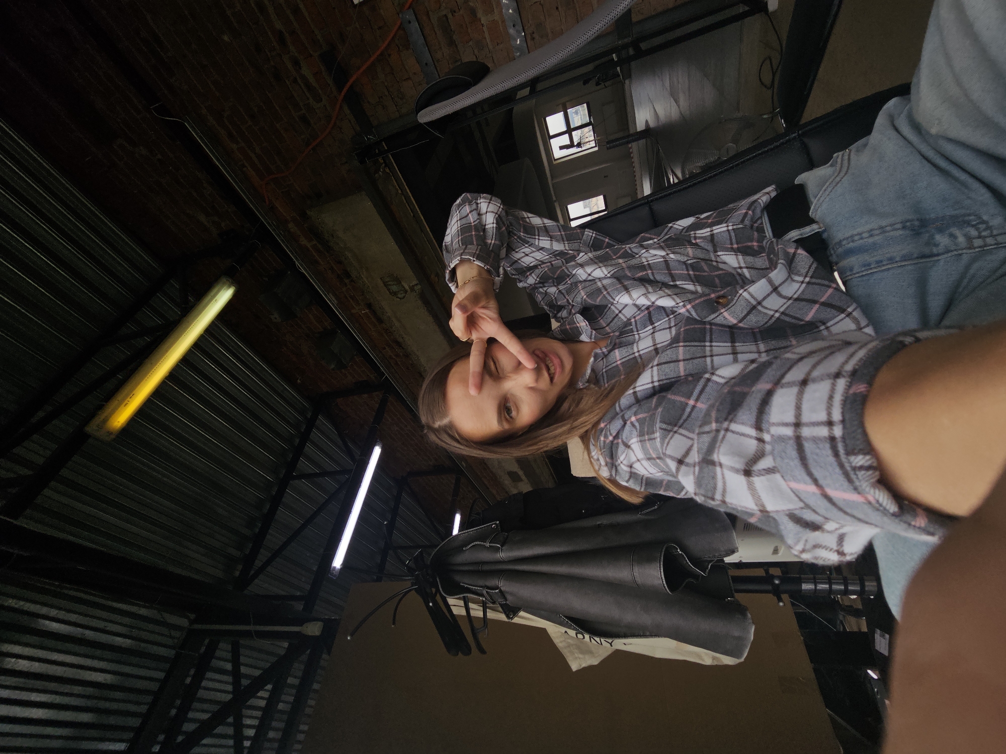 A person is holding a camera at a low angle, capturing a selfie. The individual has light brown hair and is wearing a grey and pink plaid shirt. They are making a peace sign with their fingers near their eye. The person is wearing blue denim clothing. The background includes a metal ceiling with overhead lighting. A clothes rack with garments, including a black leather jacket, is present. Based on structural features visible in the image, the photo could potentially be taken indoors in an industrial or commercial space. While it is impossible to definitively confirm the location as Chaikovsky, Russia from the image alone, the background and clothing could be consistent with an indoor environment in that city.