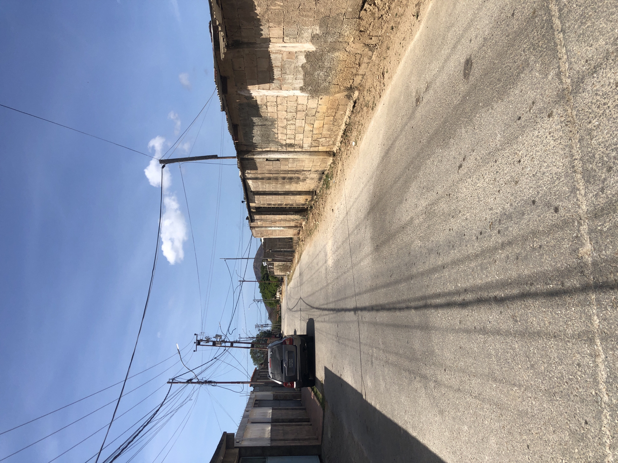 The image depicts a bright daylight scene of a narrow, paved street in Altagracia, Venezuela, looking down the road from a slightly elevated perspective. The street is asphalt, showing some wear and stains, with prominent, long shadows cast across its surface by utility poles and overhead wires, indicating a clear, sunny day.
On the right side of the street, a long, tall wall made of rough, light-colored stone blocks and concrete runs parallel to the road. Parts of the wall appear unplastered or damaged, revealing the underlying construction. On the left side, closer to the viewer, are what appear to be the tops of other buildings or walls, also suggesting a somewhat aged or utilitarian construction style. Further down the left side, a dark-colored car, possibly an SUV or hatchback, is parked facing away from the camera. Its license plate is visible but unreadable.
A dense network of utility poles and wires dominates the mid-ground and foreground on the left side, stretching across the sky. The sky above is a clear, bright blue with a few scattered, fluffy white clouds. In the distance, beyond the car and the end of the visible street, there are green trees and what appear to be hills or mountains. No specific activity or people are visible in the static scene. There is no discernible text beyond the unreadable license plate on the car.