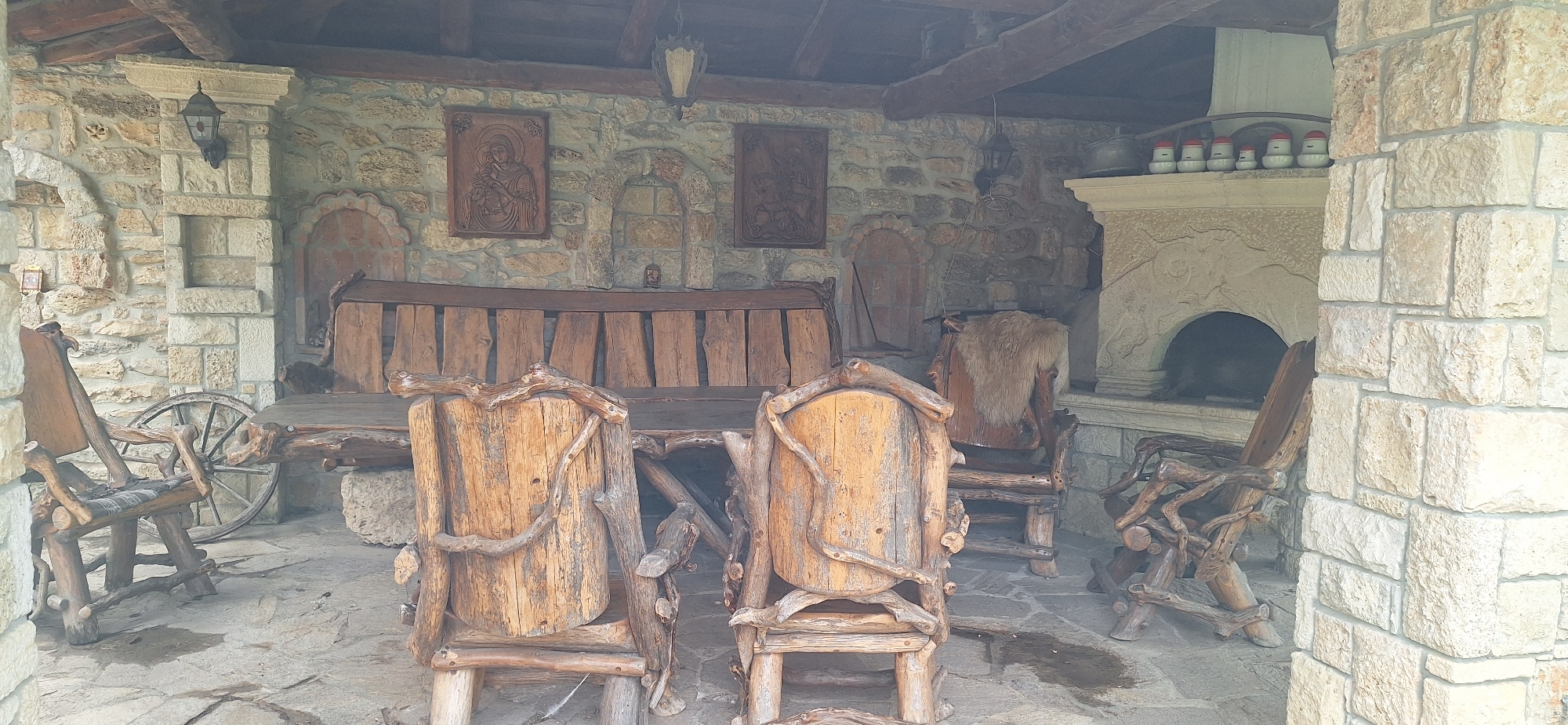 The image shows an outdoor seating area with rustic wooden furniture and a stone wall background. The furniture includes a long bench, a table, and several chairs, all made from natural wood with branches and logs incorporated into their design. Two carved wooden panels are mounted on the stone wall above the bench. To the right, there is a stone fireplace with an arched opening and decorative carvings. Several small jars with red lids are lined up on the mantelpiece. A wagon wheel is leaning against the stone wall to the left of the bench. The ground is paved with flagstones, and there are some wet patches on the ground. The overall impression is of a cozy, traditional outdoor dining or lounging space. The scene is likely located in the village of Kalimantsi, Bulgaria, given the distinctive architectural style and rustic aesthetic that is common in such locations.
