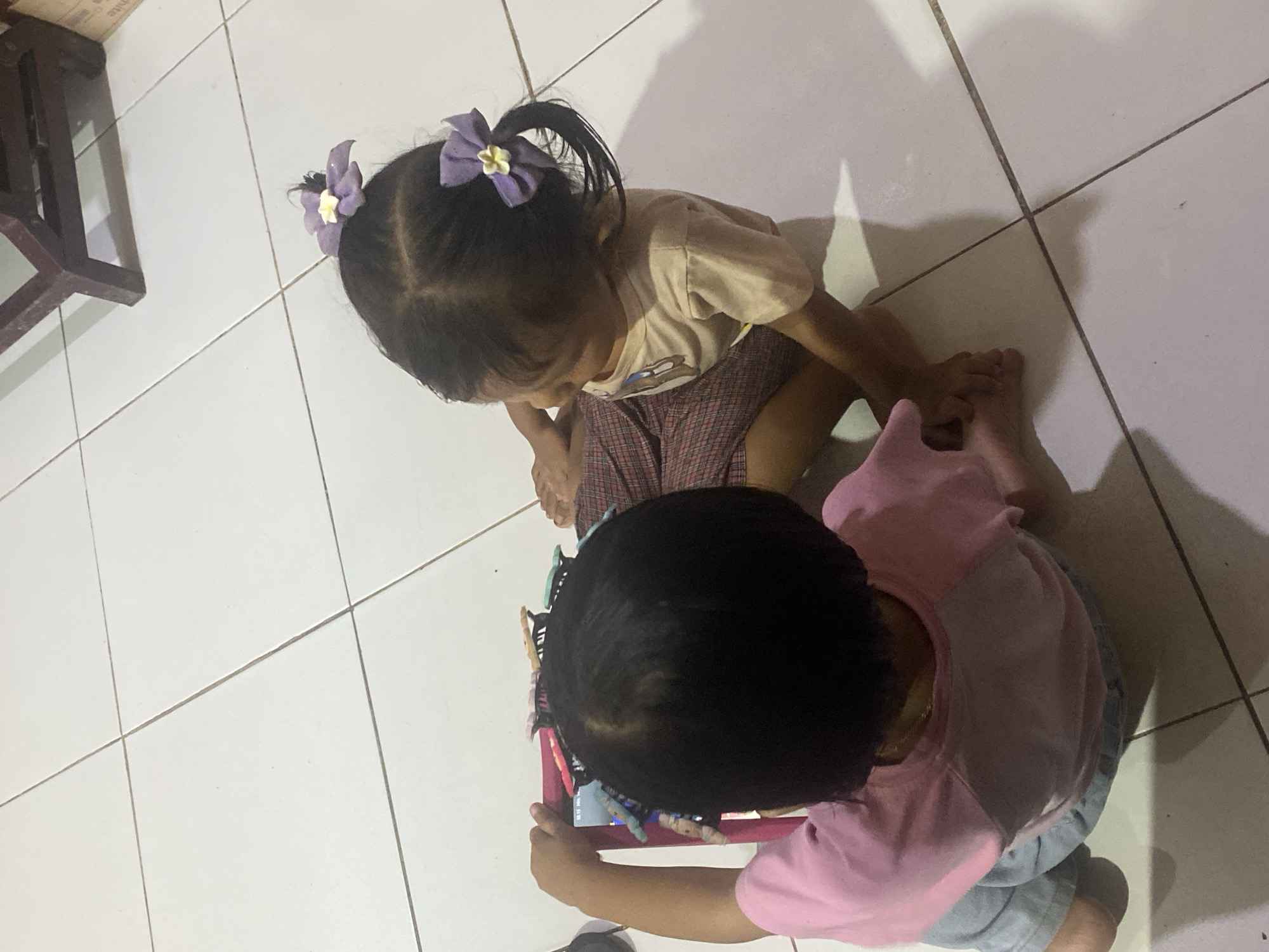 Two young children are sitting on a white tiled floor, engaged with a tablet. The child on the left, viewed from behind, is wearing a pink shirt and blue pants, and holds the tablet with both hands. The child on the right, facing away from the camera, wears a beige t-shirt and plaid shorts, and has two purple flower hair clips in their braided hair. The floor is clean and appears to be indoors, with shadows cast by the children and furniture in the background, which seems to be a wooden table or chair leg. The overall setting suggests a domestic environment in Ta', Indonesia.