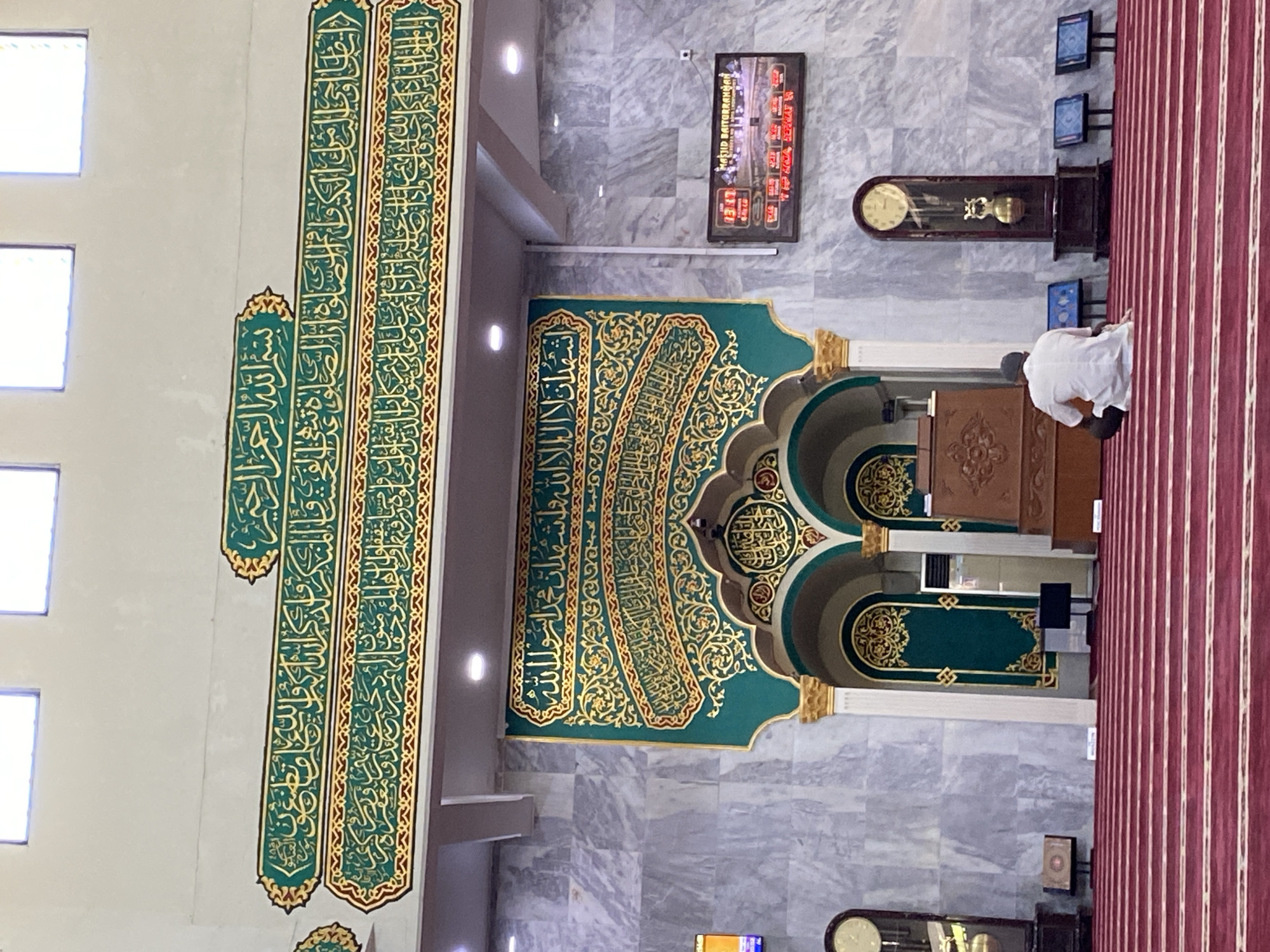 The image shows the interior of the Baiturrahman Grand Mosque in Lhokseumawe, Indonesia. The mosque features ornate Islamic calligraphy in gold against a teal background above a mihrab. A man in a white shirt is seated on the red carpeted floor, facing the mihrab. In the background, a large grandfather clock and a digital display showing prayer times are visible. The walls are covered in marble tiles.