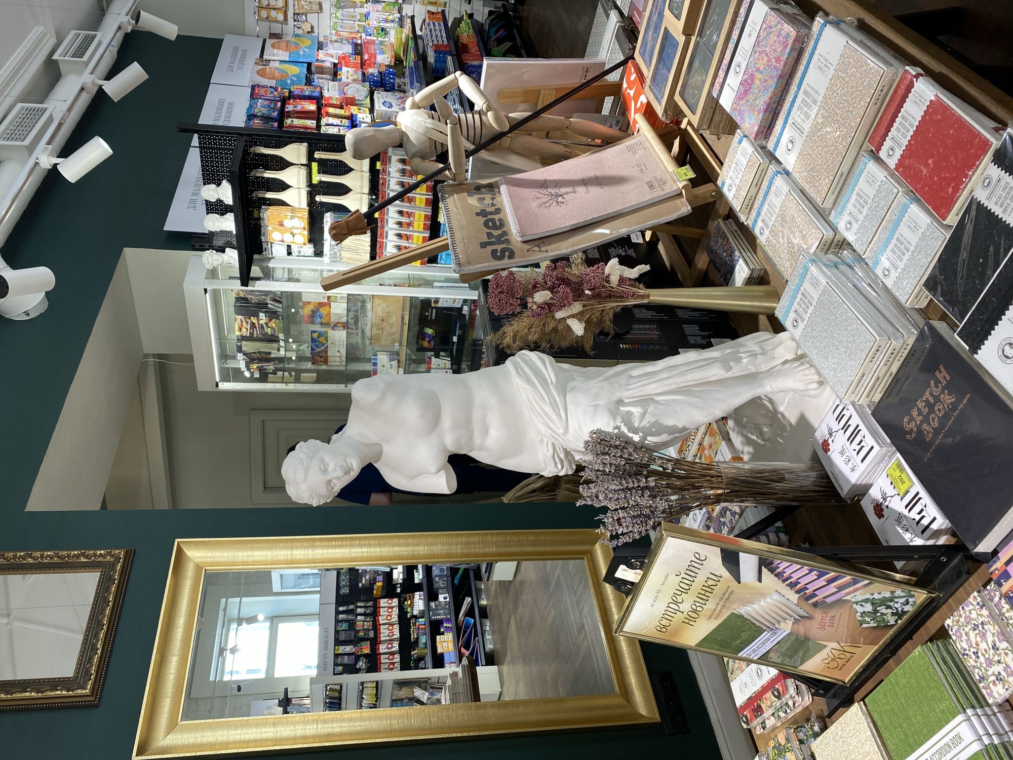 The image depicts an interior scene of an art supply or craft store, characterized by a blend of classical artistic elements and modern materials. The setting features dark green walls and bright overhead lighting.
A prominent feature is a large white plaster statue resembling a classical female figure, possibly a Venus or Ariadne, reclining on a display shelf. Next to it, a bouquet of dried lavender and other dried flowers adds a rustic touch. Nearby, a wooden art mannequin is posed with an easel or stick, surrounded by several sketchbooks. Beyond this central display, shelves are packed with a wide array of art materials, including various types of sketchbooks, notebooks, and paper in different textures and colors. Other visible items include brushes, paints, and assorted creative supplies.
Two gilt-framed mirrors are mounted on the green wall, reflecting parts of the store, including more shelves of products and a distant window or light source. The reflections also show a glimpse of a person, likely a customer or staff member, standing further back in the store. The overall impression is one of an inviting and well-stocked establishment catering to artists and crafters.
Visible text in Russian includes "ТОВАРЫ ДЛЯ ТВОРЧЕСТВА" (Goods for Creativity) on a banner, "ДЛЯ МАЛЕНЬКИХ ХУДОЖНИКОВ" (For Young Artists) on another section, and "встречайте новинки" (Meet the New Arrivals) with "пополнение коллекции блокнотов Potentate" (Replenishment of the Potentate Notebook Collection) on a display sign. English text includes "SKETCH Book," "If at first you don't succeed, try, try again." on a sketchbook cover, "DECKLE EDGED WATERCOLOR PAPER," and brand names like "POTENTATE" and "SM-LT." Chinese characters "水彩纸" (Watercolor paper) are also visible.