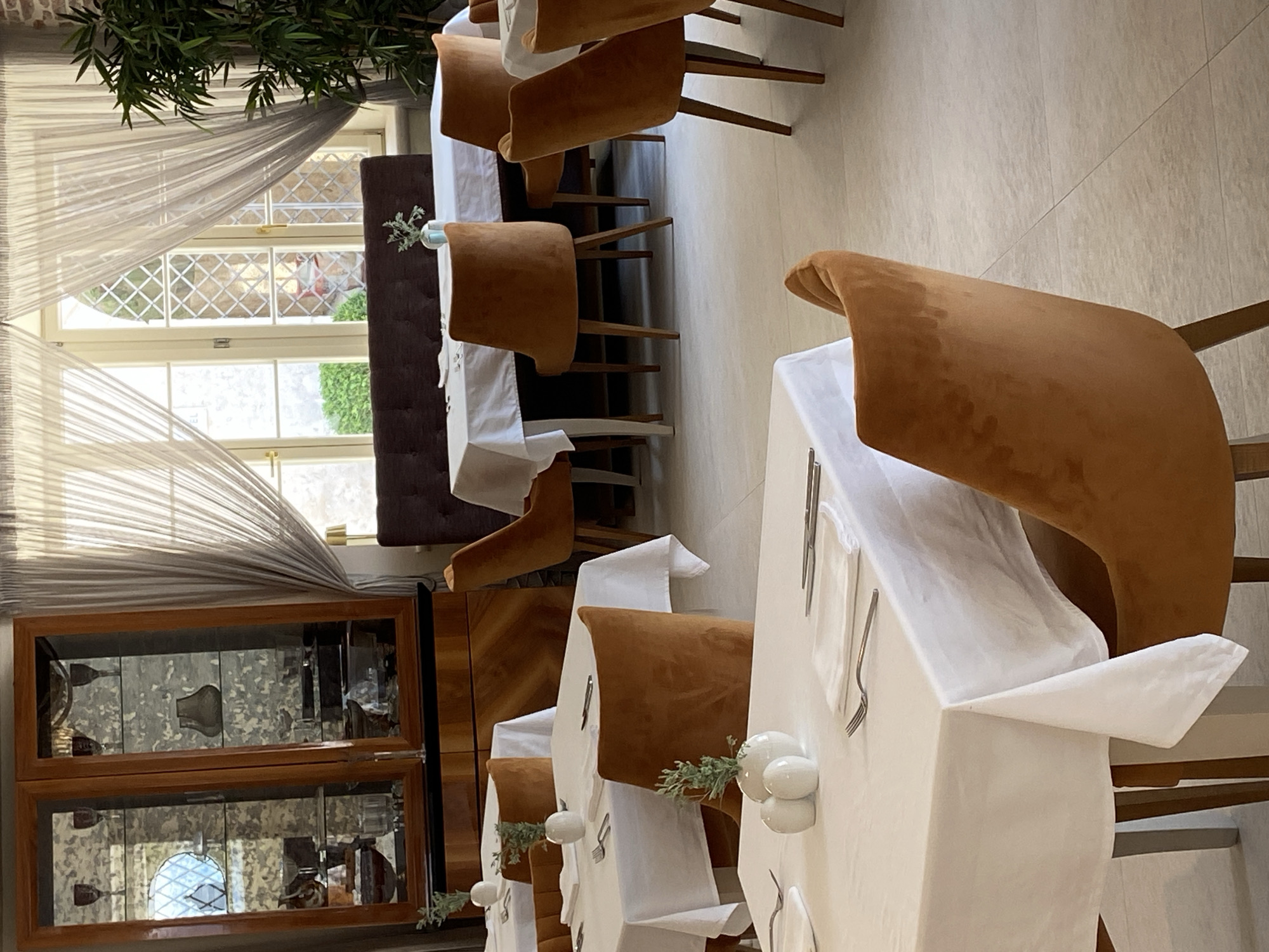 The image depicts the interior of a restaurant in Perast, Montenegro, with a focus on the dining area. Several tables are set with white tablecloths, white napkins, silverware, and small decorative vases with greenery. The chairs are upholstered in a warm, reddish-brown velvet fabric. In the background, a large wooden cabinet with glass doors displays various glassware and decorative items. Large windows with sheer curtains allow natural light to enter the room. A tall potted plant is visible on the right side of the image. The flooring appears to be light-colored tile. The scene is staged for dining, but no people are present.