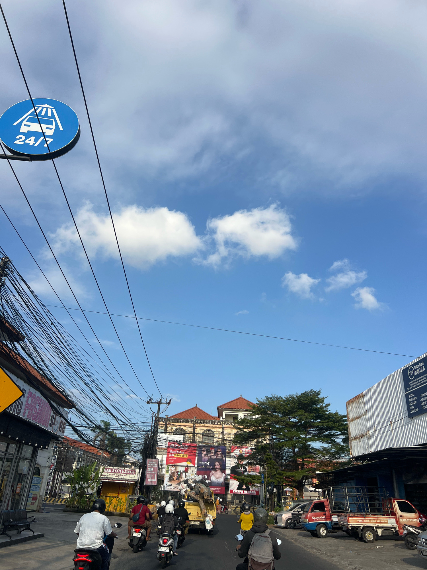 A street scene in Denpasar, Indonesia, features a clear blue sky with scattered clouds. A circular blue sign with a white graphic of a car being washed and the text "24/7" is visible in the upper left quadrant, suspended above a tangled mass of overhead electrical wires. The street is lined with buildings of various architectural styles, including a traditional building with a terracotta roof and an arched facade, and a more modern building with a corrugated metal exterior. Several shops are visible, with signage in Indonesian, such as "FASHION STORE" and "NASI AYAM HAINAN". A yellow food stall is also present. Traffic on the street includes several motorcycles with riders wearing helmets and a yellow truck with a load of wood. A white car and a pink car are parked on the side of the road. The overall impression is that of a busy urban street in a tropical climate.