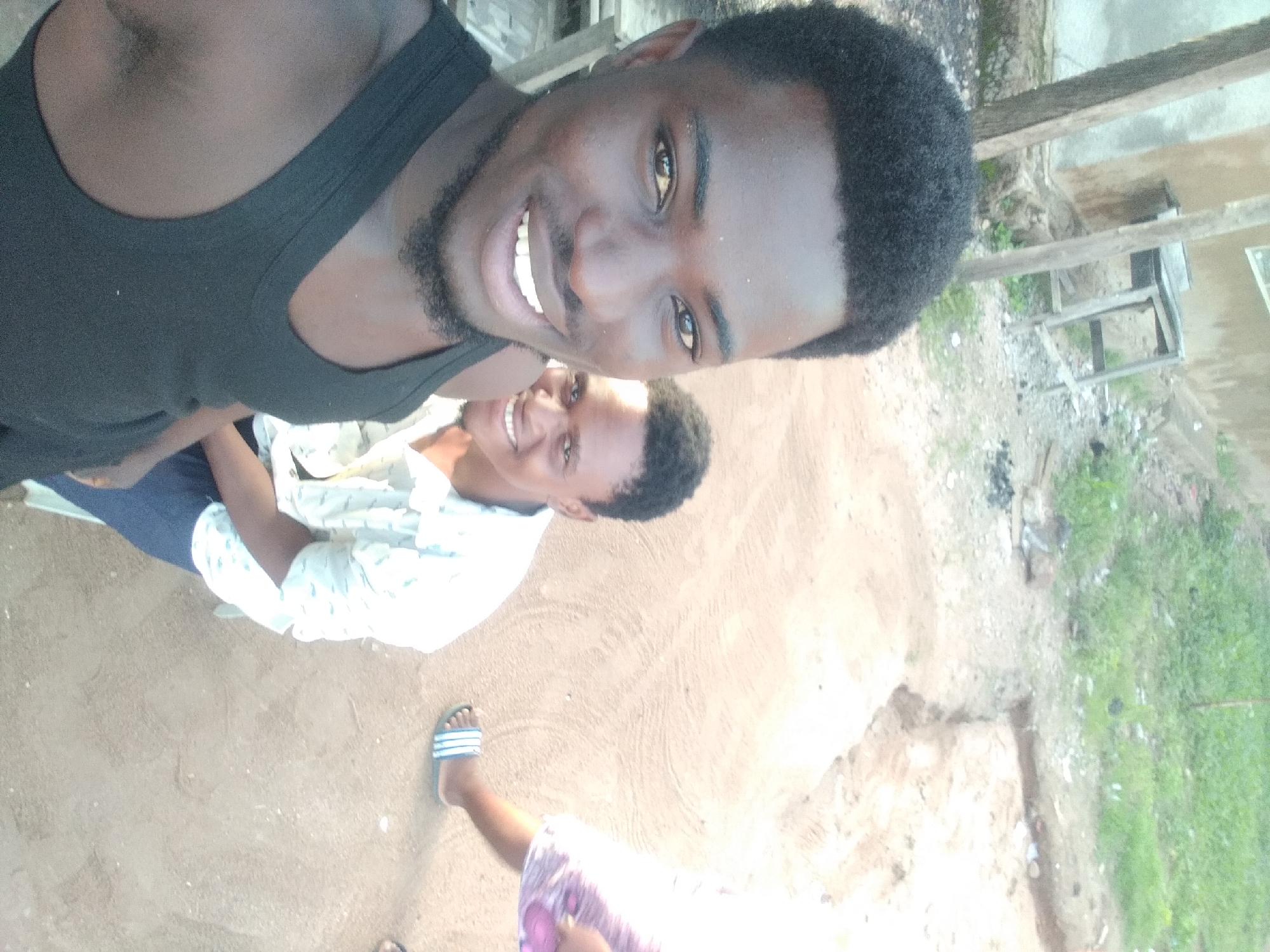 In Jalingo, Nigeria, two young Black men are captured in a selfie. The man in the foreground, on the left, is wearing a black tank top and has a short beard. He is smiling directly at the camera. Behind him, slightly to the right, is another young Black man wearing a light-colored shirt with a pattern. He is also smiling. They are outdoors in a sandy, somewhat unkempt area with scattered greenery and debris. In the background, a building with a wooden structure and a table can be seen. The lighting suggests it is daytime, possibly late afternoon.