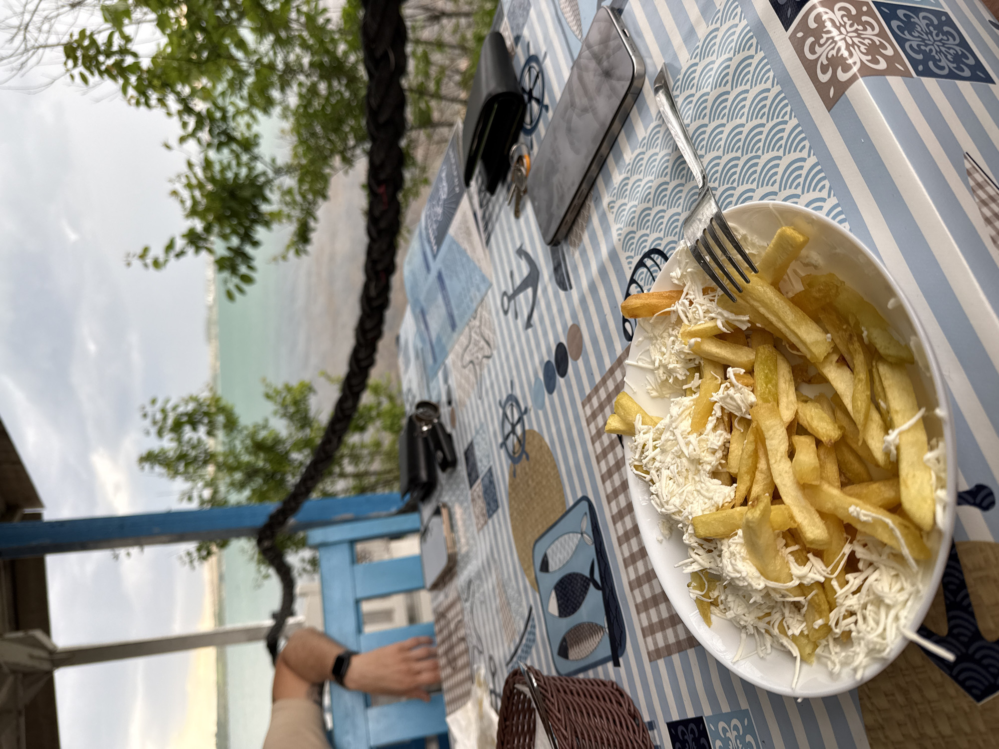 The image shows a plate of french fries topped with shredded cheese on a table with a blue and white nautical-themed tablecloth. A fork rests on the edge of the plate. Beside the plate, on the table, are a smartphone, a wallet, and keys. In the background, visible through the open-air structure, is a blurred view of the sea and trees. A blue wooden structure and a braided rope are also visible. A person's arm and wrist with a watch are visible in the lower left corner. The image was likely taken in a restaurant or cafe located in Burgas, Bulgaria, given the sea view and typical seaside restaurant setting.