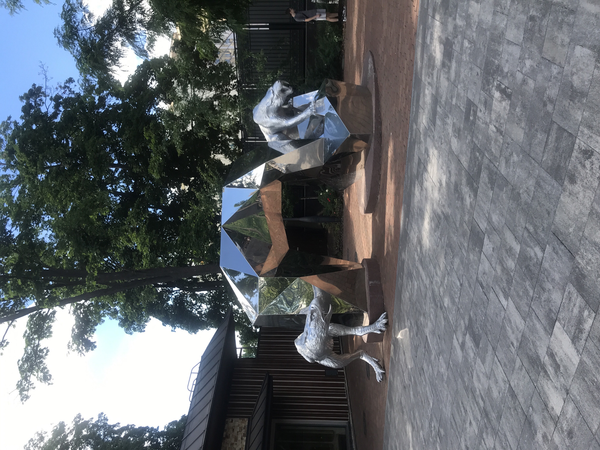 In Kharkiv, Ukraine, an outdoor sculpture depicts two stylized metallic animals emerging from a large, faceted, crystalline structure. On the left, a silver creature with sharp claws and an eagle-like head is positioned as if stepping out from behind the crystal. On the right, a silver saber-toothed cat is crouched within a recess of the crystal. The sculpture is situated on a reddish-brown brick pathway, which is bordered by a paved area with grey interlocking stones in the foreground. Behind the sculpture, a modern building with dark siding and a metal roof is visible. Lush green trees with dense foliage line the background, with a clear blue sky overhead. A man in a t-shirt and shorts is standing to the right of the sculpture, observing the scene. The overall setting appears to be a public space or park area.