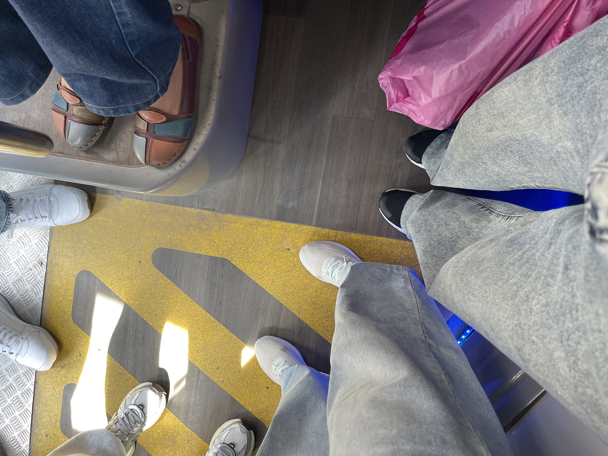 The image shows a view of the floor of what appears to be a public transportation vehicle, possibly a train or bus. The floor has a yellow and gray striped pattern, with a metal grate section visible on the left. Several pairs of feet and lower legs of different people are visible. One person is wearing blue jeans and brown and blue patchwork-style loafers. Another person is wearing light-wash jeans with black and white sneakers and carrying a large pink plastic bag. A third person is wearing light gray or white sneakers with light-wash jeans. A fourth person is wearing white sneakers. The angle of the image is from above, looking down at the floor. The setting seems to be indoors, likely inside a moving vehicle, in the city of Unknown, Unknown.