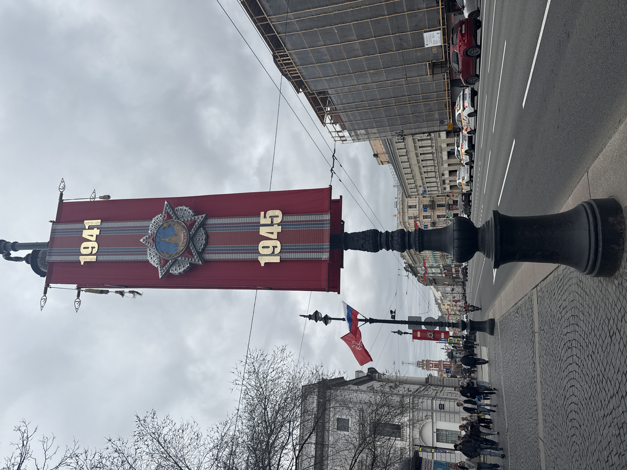 The image captures a street scene in Saint Petersburg, Russia. The central focus is a decorative lamppost adorned with a large, red banner bearing the golden numbers "1941" and "1945," flanking a star-shaped emblem. This commemorates the Great Patriotic War (World War II). The lamppost stands on a cobblestone sidewalk. A line of people is walking down the sidewalk. In the background, another lamppost bears flags, which appear to be Russian. Along the street are cars parked along the side of the road. Buildings line the street on both sides. The sky is overcast. The general atmosphere suggests a commemoration or event related to World War II.