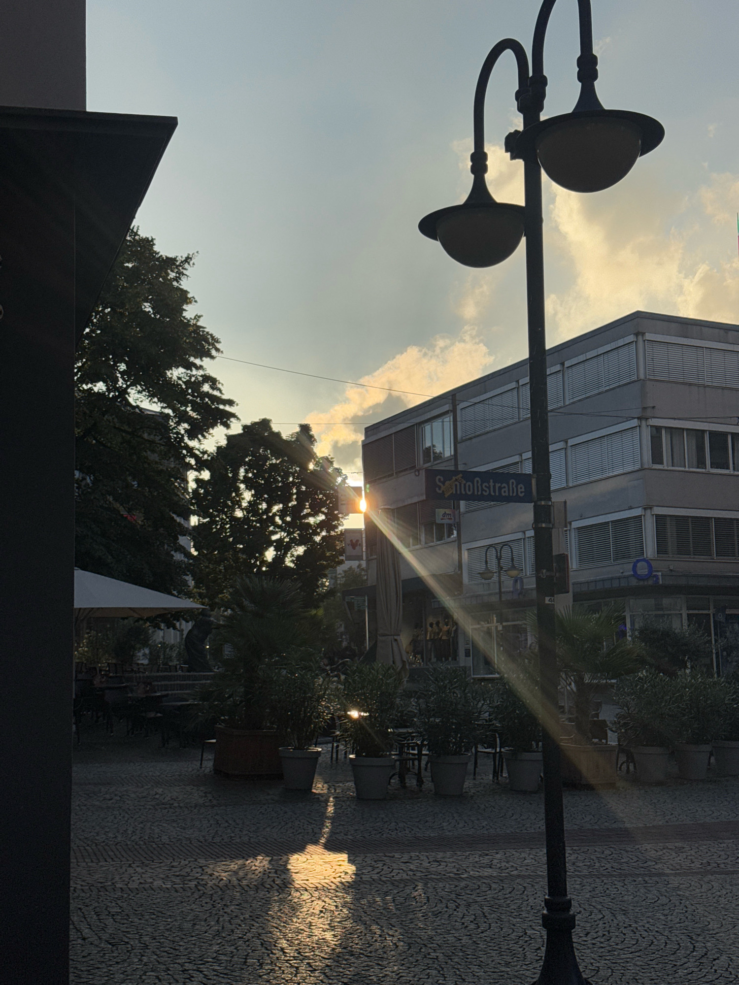The image captures a street scene in Cleves, Germany, during sunset. A prominent black lamppost with two lamps stands in the foreground, casting a dramatic lens flare across the frame. To the left, a dark building corner is visible, with trees and a white awning indicating an outdoor seating area for a cafe or restaurant.
In the background, a modern, multi-story building with numerous windows and horizontal blinds dominates the skyline. A street sign on a pole reads "Schloßstraße," identifying the location. The sky is partly cloudy, with the setting sun filtering through the clouds and trees, creating a warm, golden light. The ground is paved with cobblestones, and several potted plants are arranged in front of the building, some of which are illuminated by the low sunlight. The overall impression is a peaceful urban environment bathed in the soft light of the golden hour. No people are clearly visible in the scene.