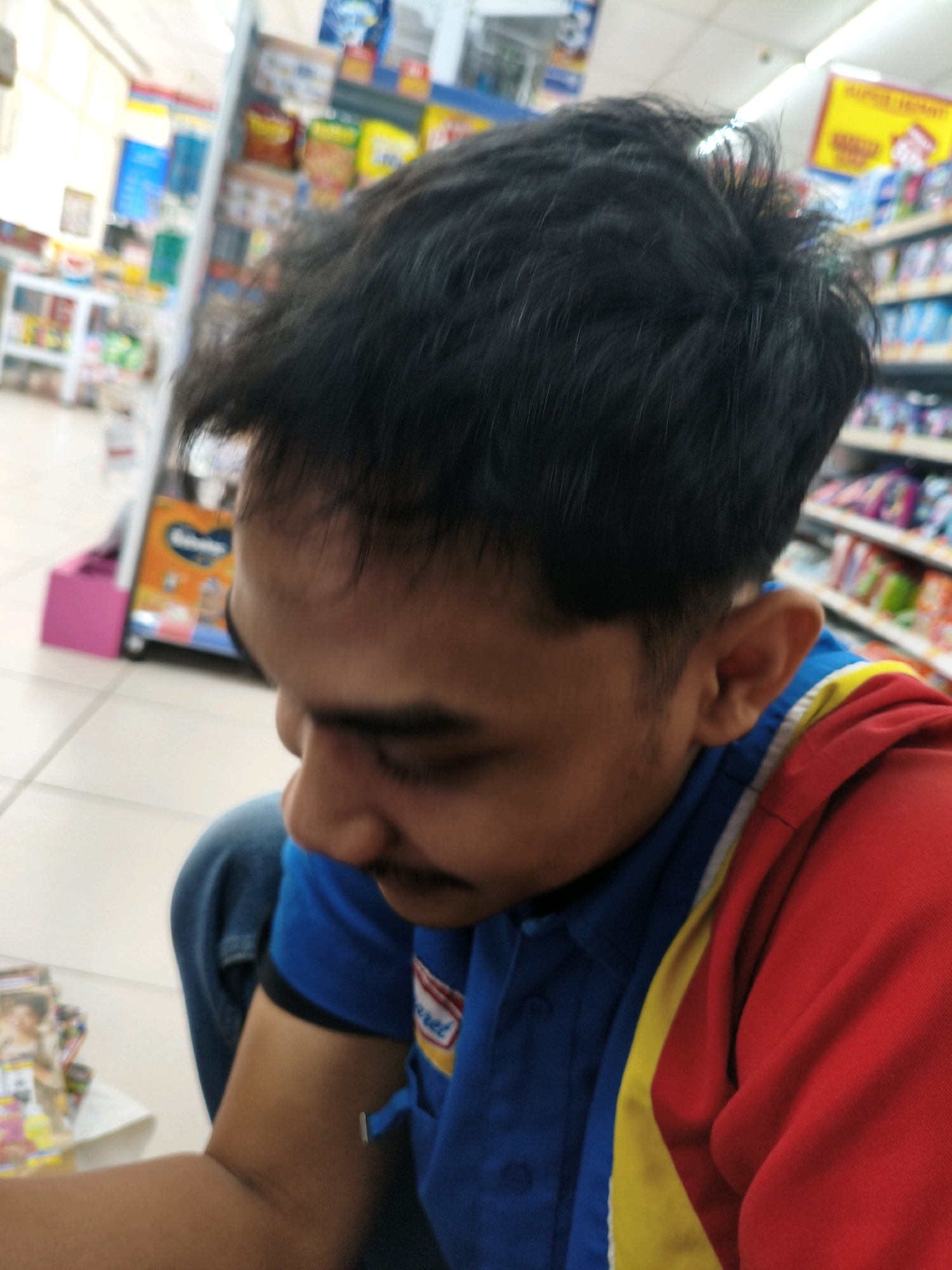 The image shows a close-up of a young man's head and shoulders, taken from a low angle. He has dark, messy hair and is wearing a blue shirt with red and yellow accents. He is looking down, with his face partially obscured. The background is blurred but appears to be an indoor setting with shelves stocked with various products, suggesting a retail environment like a minimarket. The lighting is bright and appears to be artificial. The man's posture and the blurred background suggest he might be in the process of taking a selfie or being photographed in his place of work. The specific location of Sumberejo Kulon, Indonesia, is not visually evident from the image itself, but it is provided as context.