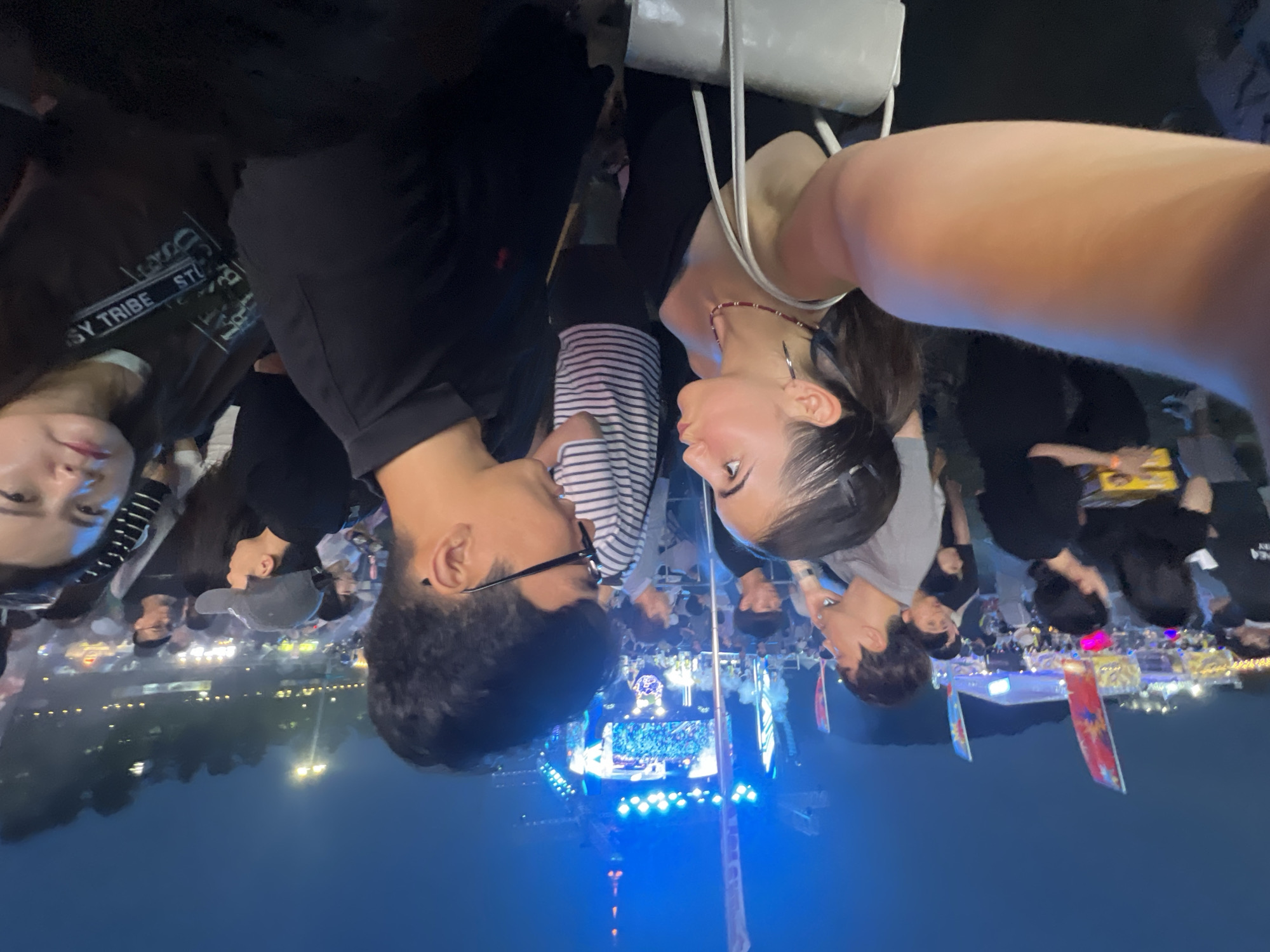 This is a candid, eye-level, wide-angle photograph taken at night during an outdoor event, possibly a festival or concert, at the Daegu Stadium in Daegu, South Korea. The foreground is dominated by a couple facing each other in an intimate moment, with the camera seemingly held by one of them. The woman on the left, with dark hair pulled back in a ponytail, is wearing a black sleeveless top and a necklace, and has a white purse. The man on the right, with dark hair and glasses, is wearing a black polo shirt.
Behind the couple, a large crowd of people is visible, suggesting a well-attended event. In the background, there is a brightly lit stage with large screens displaying imagery, and tall structures, possibly part of the stage setup or surrounding city. Various colorful banners and flags are also present, adding to the festive atmosphere. The sky is dark, indicating it is nighttime, with some lights from the event and surrounding areas visible. The overall environment is bustling and energetic, characteristic of a public gathering.