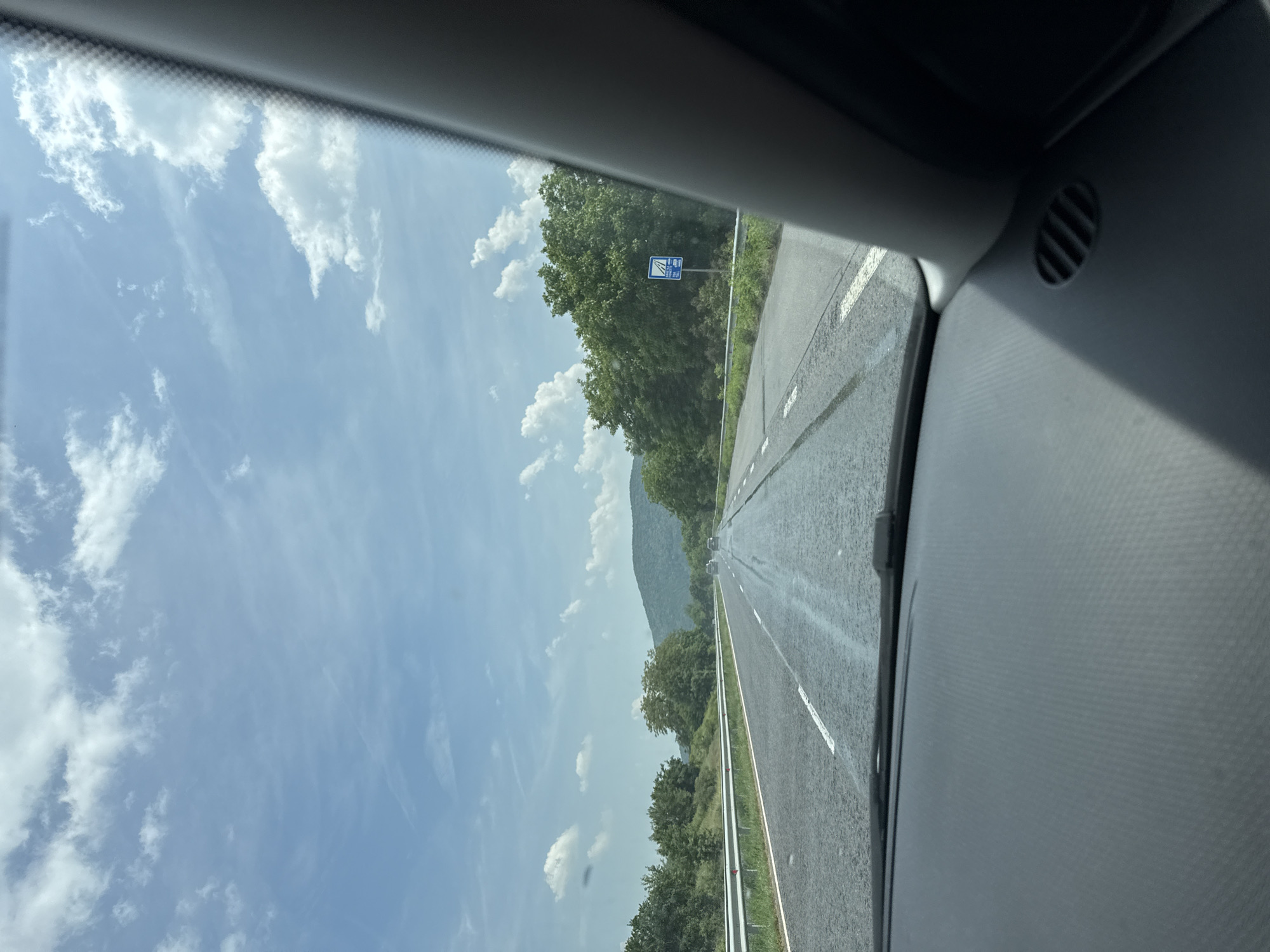 The image depicts a straight section of a two-lane asphalt road (one lane per direction) viewed from inside a moving vehicle, specifically in the city of Zmeyovo, Bulgaria. The car's dark gray textured dashboard, windshield wipers, and a circular air vent on the right side are visible in the foreground.
The road surface appears damp or wet in various patches, with visible tire streaks or marks on the asphalt. White solid and dashed lane markings are present. Metal guardrails line both sides of the road.
Beyond the guardrails, dense green trees and bushes form a natural barrier, extending into the midground and background. In the distance, several vehicles are visible traveling in the same direction on the road. A prominent blue rectangular traffic sign, featuring a white pictogram of a road with a barrier and faintly visible text including "TOLL," is positioned on the right side of the road.
The background features a range of tree-covered hills or mountains under a partly cloudy sky. The sky is bright blue with scattered white cumulus clouds, along with some higher, thinner cirrus clouds. No people are discernible in the scene.