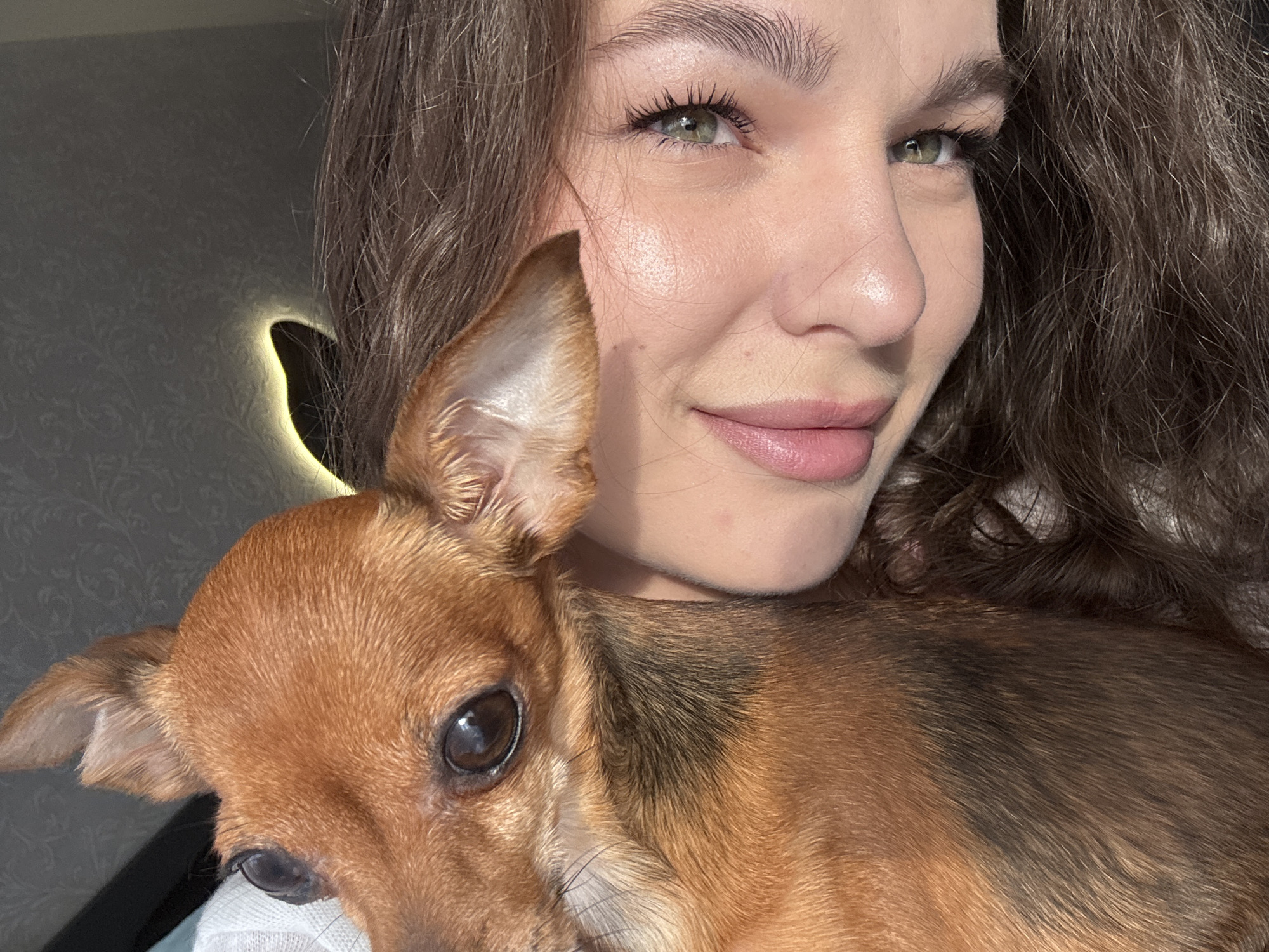 A woman is posing for a selfie with a small, brown, short-haired dog. The dog is positioned close to her face, partially obscuring her body. The woman has long, wavy brown hair, green eyes, and light skin. She is smiling slightly. In the background, there is a wall with a decorative, illuminated object resembling a crescent shape. The location is Chaikovsky, Russia.