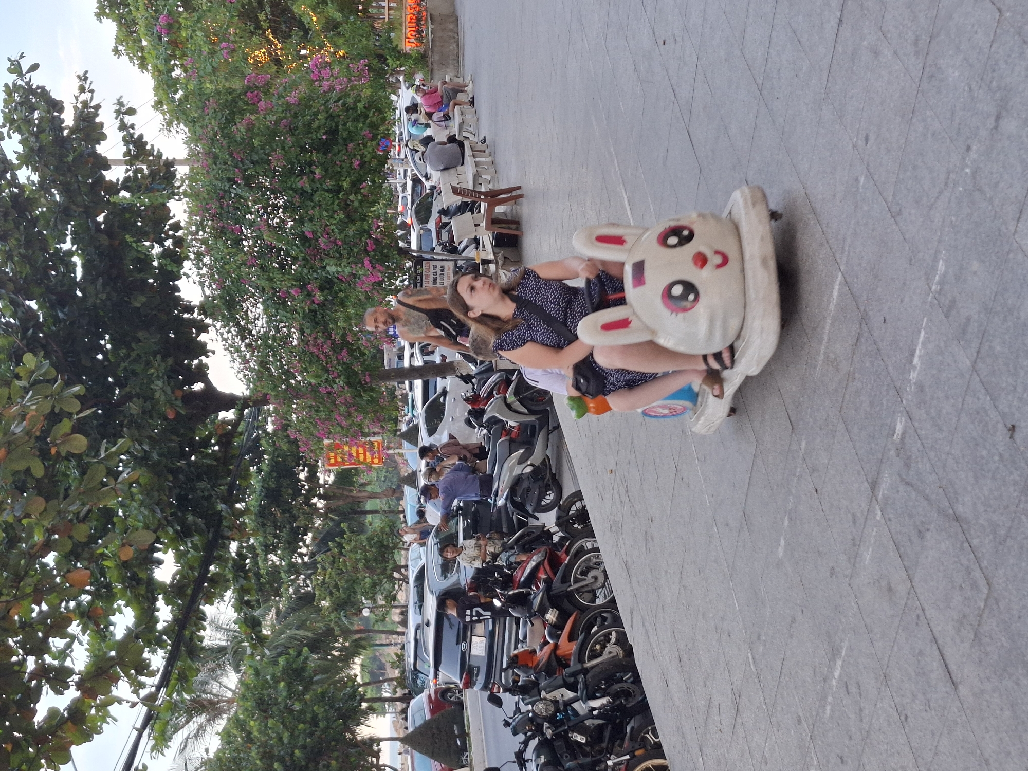 In a street scene in Bac Nha Trang Ward, Vietnam, a woman is riding a small, bunny-shaped toy car on a paved sidewalk. She is wearing a dark blue, patterned dress and sandals. Her legs are visible from the knees down. A child, partially obscured, is sitting behind her. The toy car is white with pink accents, featuring large eyes and a red nose on the front. Behind them, a man with a muscular build and tattoos is standing, wearing a dark bag slung over his shoulder. Further in the background, several motorbikes are parked, and beyond them, cars are visible on a street lined with trees and flowering bushes. The environment suggests a public space, possibly a promenade or a busy street.