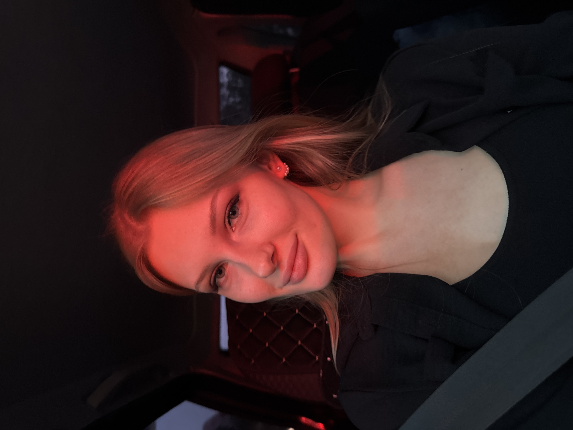 A young woman with blond hair is shown in the photograph. She is positioned inside a car, as indicated by the seatbelt visible across her chest and shoulder. The background is dark, with some red lighting. The quilted texture of the car's interior is partially visible behind her. The photograph seems to have been taken inside a car at night. There is no evidence to pinpoint the location as Perm, Russia.