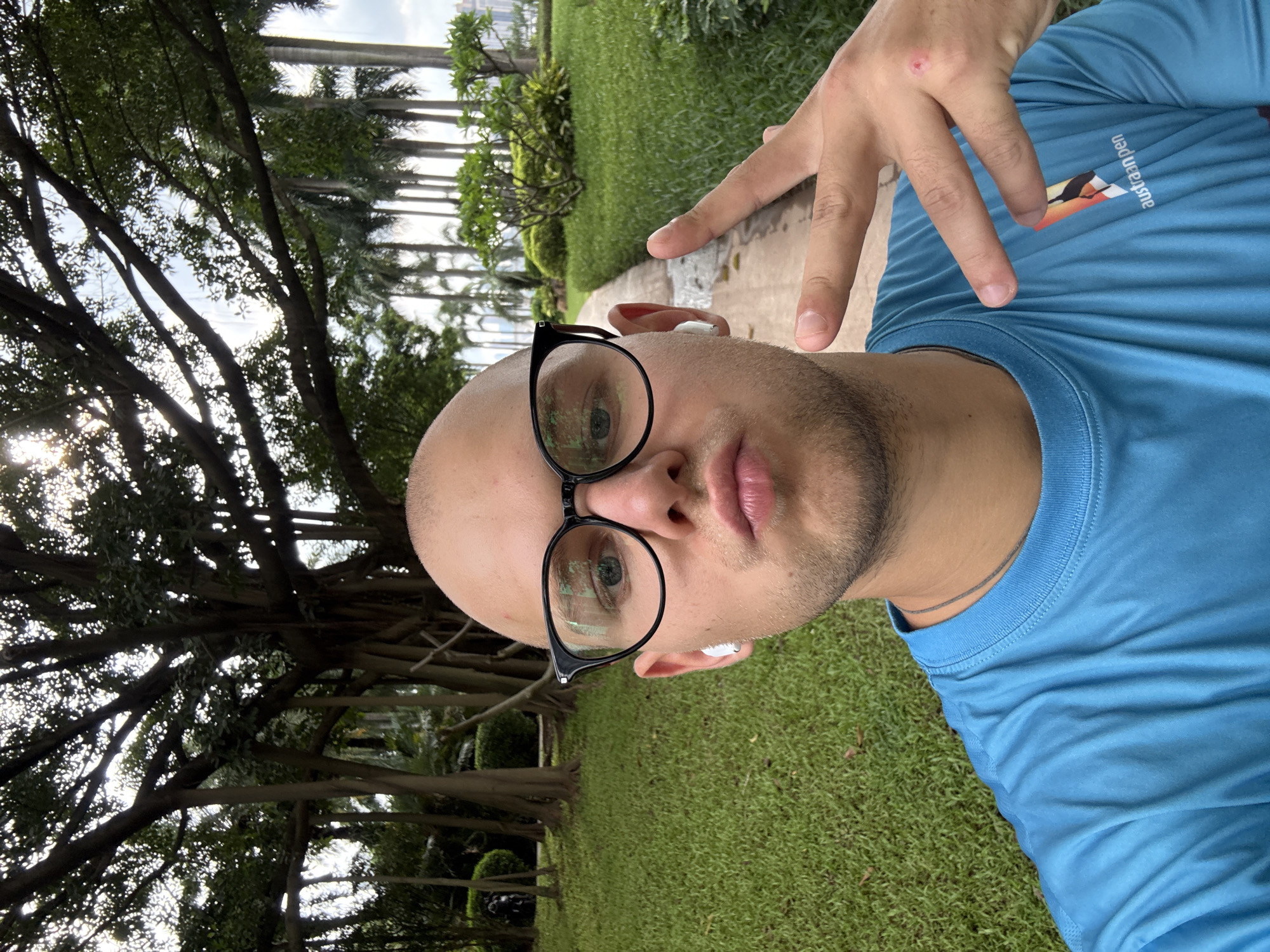 A bald, fair-skinned man in his 20s wearing black-rimmed glasses, white earbuds, and a bright blue t-shirt is taking a selfie. He is outdoors, with a path and lush green grass behind him. Tall palm trees and a large, mature tree with aerial roots are visible in the background. The man is holding up two fingers in a peace sign with his right hand, and his lips are pursed. A logo with a bird and the text "austraanpen" is visible on the right side of his t-shirt. The setting appears to be a park or garden within Haizhu District, China.