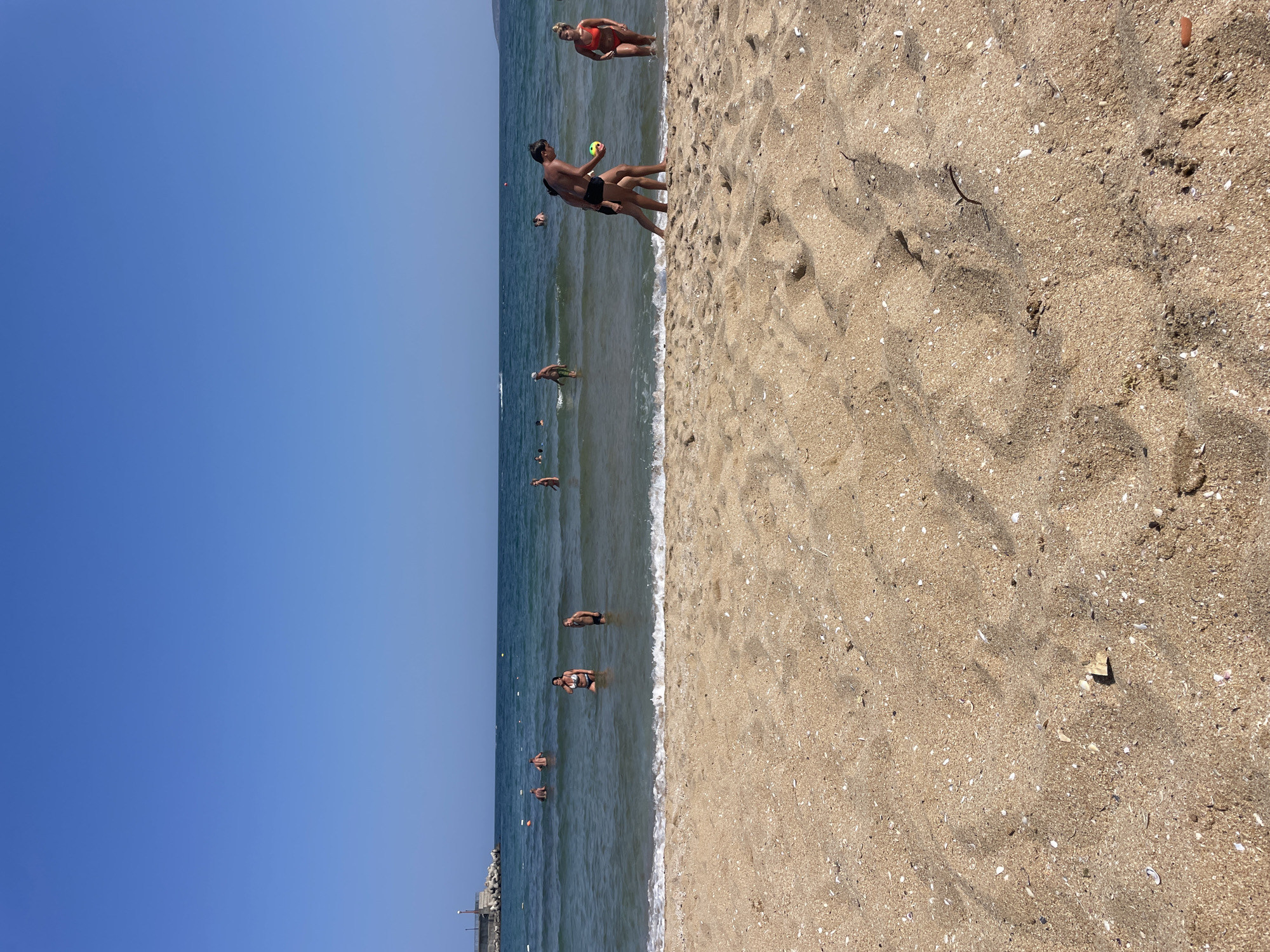 This is a photograph of a beach in Dyuni, Bulgaria, on a clear, sunny day. The foreground is dominated by a sandy beach with scattered seashells. In the middle ground, several people are in the shallow water, some wading and others swimming. The water is clear and appears calm, with small waves lapping at the shore. In the background, the ocean extends to the horizon under a bright blue sky. A breakwater structure and some distant landforms are visible on the left and right respectively. The overall scene depicts a leisurely day at the seaside.