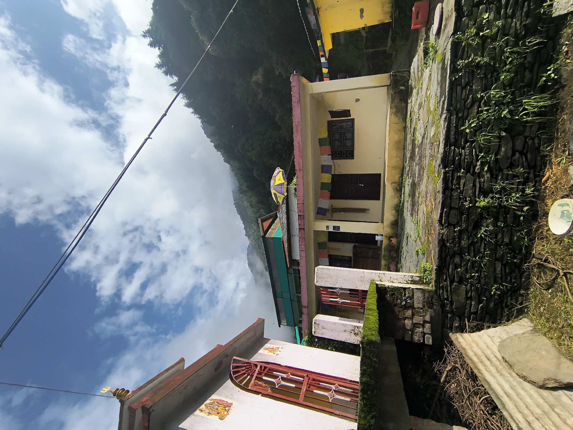 In Haini, India, a low-angle, wide-shot captures a cluster of houses nestled against a verdant hillside under a partly cloudy sky. To the left, a white building with red trim and a decorative golden spire features a red metal gate and vibrant murals on its facade. Adjacent to it, a cream-colored house with a pink roofline displays a string of colorful prayer flags. Another yellow structure, partially visible to the right, also has prayer flags. The ground in front of the houses is paved with stone, with patches of moss. A stone wall runs along the foreground, covered in green vegetation. In the background, a densely forested mountain rises, partially obscured by mist, with power lines crisscrossing the sky.