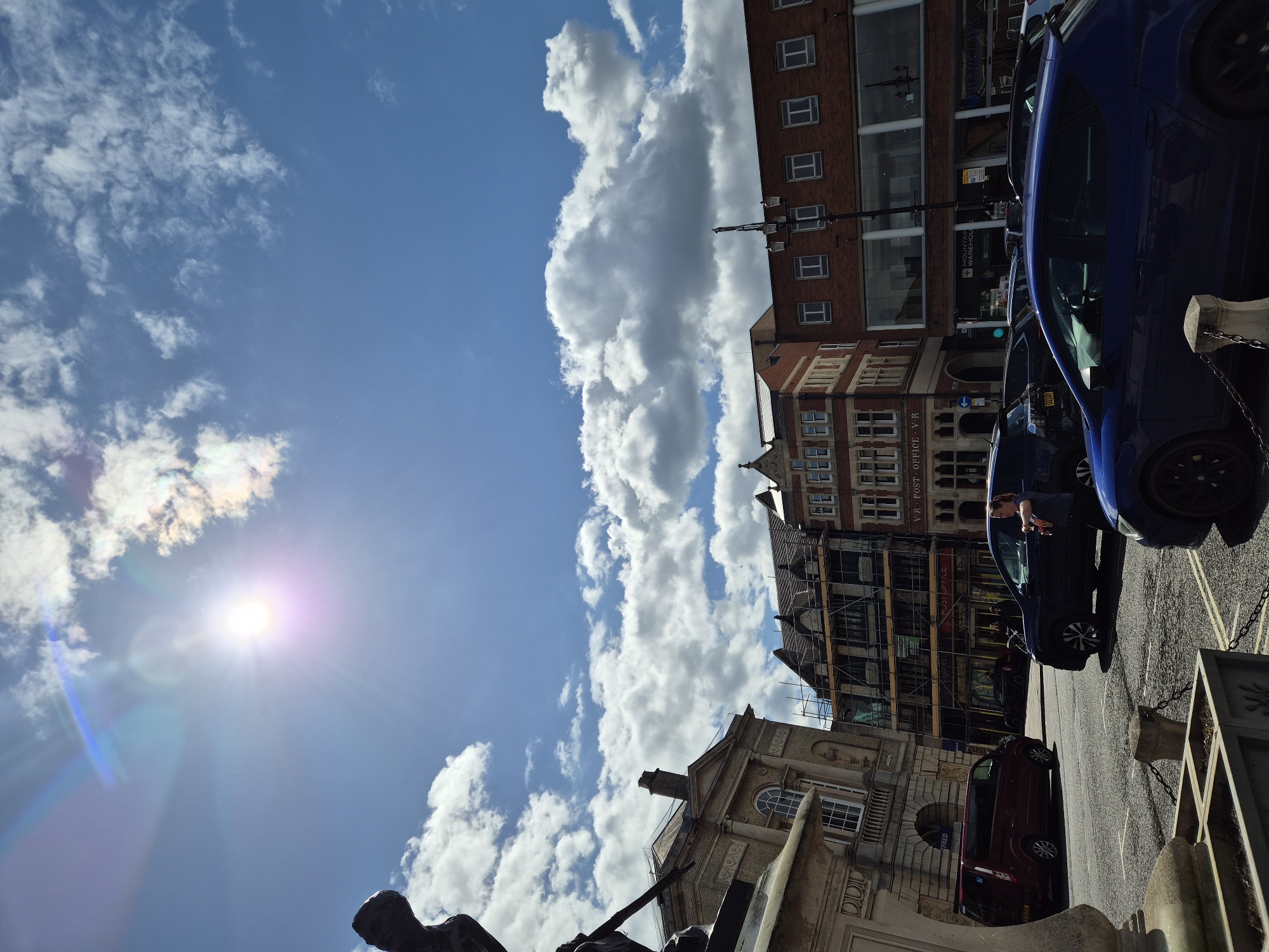 Here is a detailed description of the image:
The image is taken from a low angle, looking upwards towards a street scene in West Suffolk, United Kingdom, on a sunny day. The sky is predominantly blue with scattered cumulus clouds. The sun is visible, creating a lens flare.
The photograph captures several buildings with varying architectural styles. Some buildings feature brick facades with rows of windows, while others have ornate stonework and scaffolding, possibly indicating renovation or construction.
On the right side of the frame, several cars are parked along the street. A blue car is prominently visible in the foreground, partially obscuring the buildings behind it. A maroon-colored van is parked behind the blue car. People can be seen inside the parked cars.
In the foreground, the top part of a statue or monument is visible, suggesting the image was taken near a public square or memorial. The statue appears to be made of a dark material.