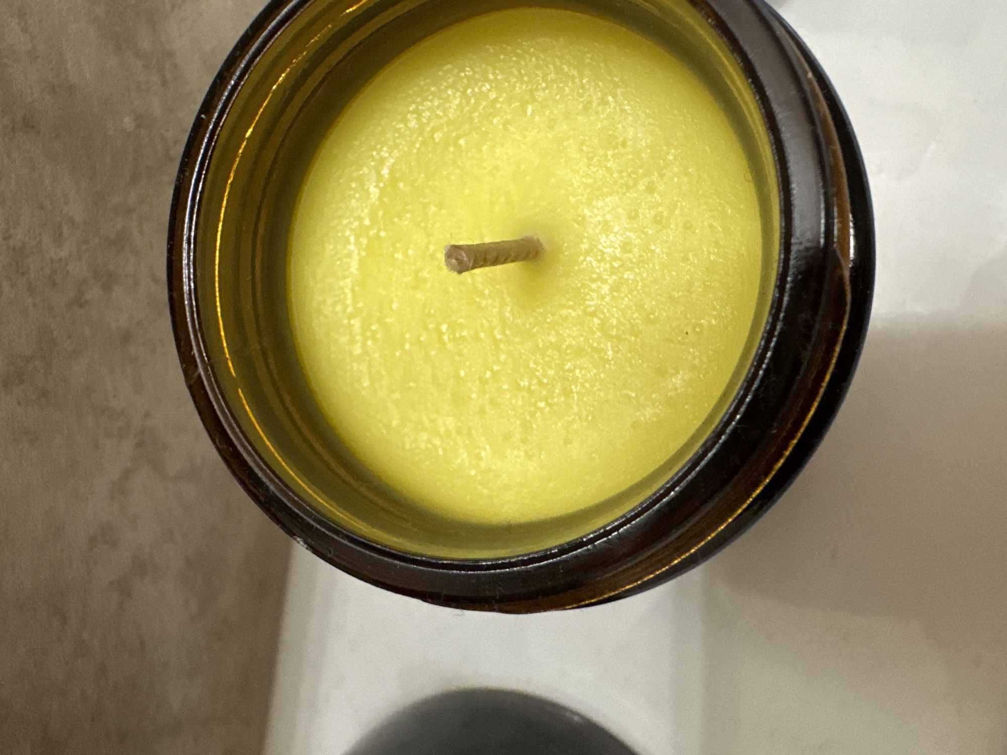 An overhead, close-up shot shows a yellow candle in a dark brown glass jar. The candle wax is textured and appears smooth, with a single wick centrally located. The jar sits on a white surface, and a blurred dark object is visible to the left. A beige, textured surface is in the background. The scene is indoors and well-lit. The specific location in Saint Petersburg, Russia, is not discernible from the image.