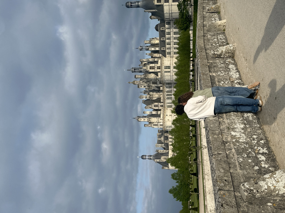 The image depicts a couple standing on a stone bridge in front of the Château de Chambord, located in Chambord, France, at coordinates 47.616 latitude, 1.514 longitude. The couple is facing the castle, with the man on the left, wearing a white jacket and blue jeans, and the woman on the right, wearing a brown shirt and blue jeans. The castle is a large, white structure with many towers and spires. The sky is cloudy with a slight grey tinge. The ground around the bridge is covered in gravel. A low stone wall lines the bridge. The bridge is covered in moss and is surrounded by trees. The setting appears to be a sunny day with a slight breeze. The scene suggests a romantic and picturesque location.