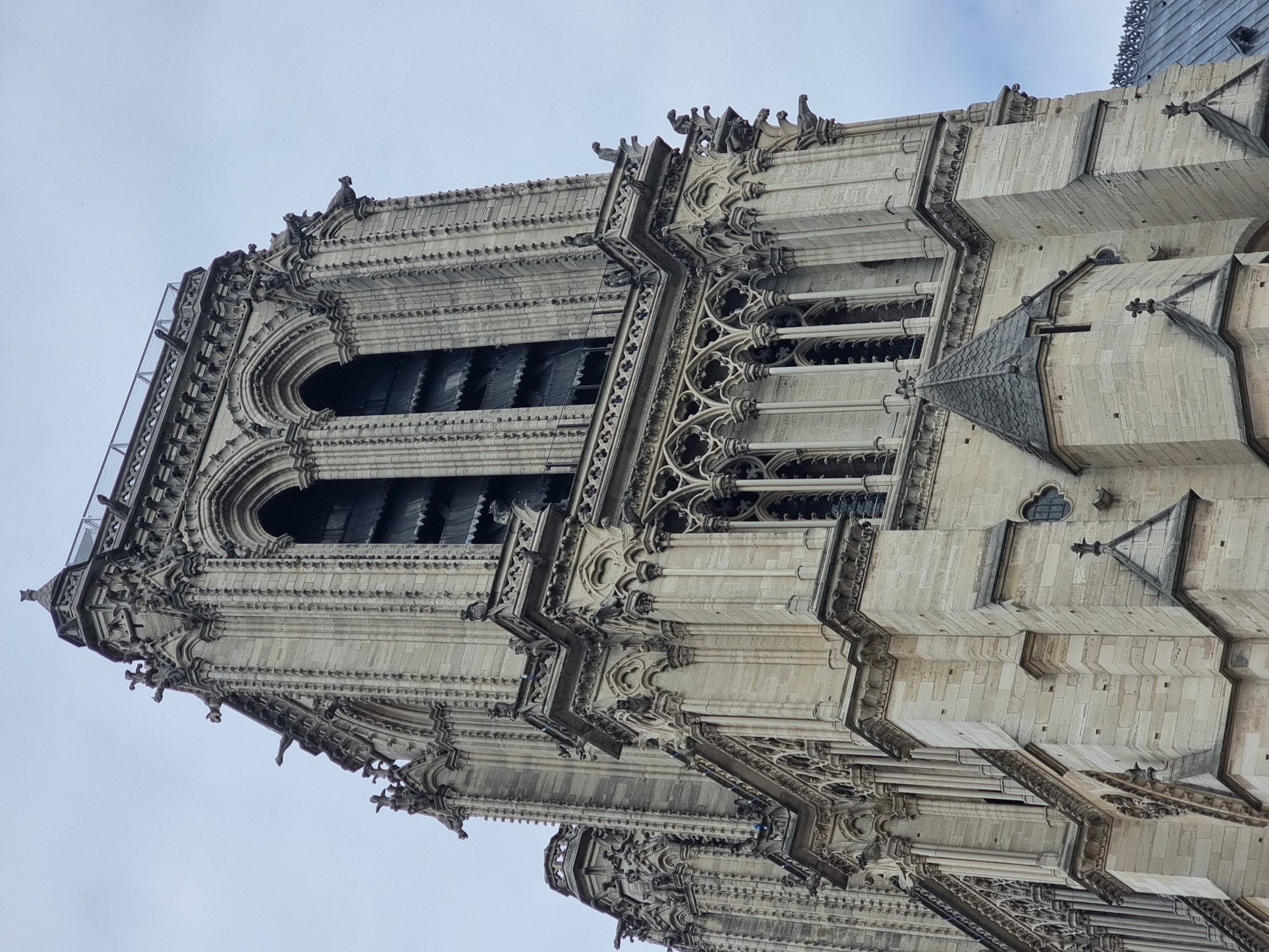 The image shows the north tower of Notre-Dame Cathedral in Paris, France. The tower is made of light-colored stone and is richly decorated with gothic architectural details, including gargoyles and pointed arches. The windows of the tower are visible, and a portion of the roof and upper walls of the cathedral can be seen on the right side of the frame. The sky is overcast.