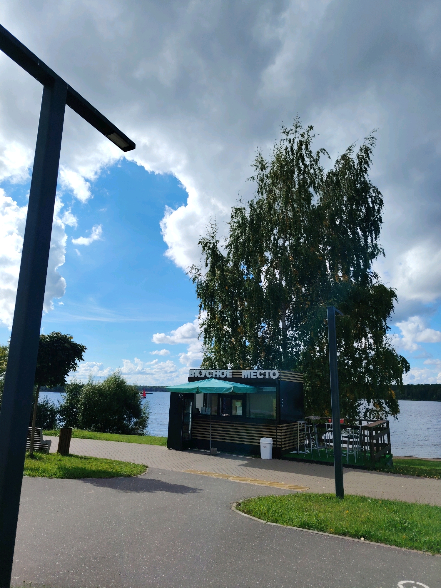This image captures a sunny day with scattered clouds at a waterfront location in Konakovo, Russia. A modern-looking food kiosk with the Russian text "ВКУСНОЕ МЕСТО" (which translates to "Delicious Place") is prominently featured. The kiosk has a green awning and appears to be open for business. Adjacent to the kiosk, there is an outdoor seating area with tables and chairs.
In the foreground, a paved walkway curves through a grassy area, and a dark, modern lamppost stands to the left. A wooden bench and a trash bin are also visible along the walkway. The background is dominated by a large body of water, likely the Volga River or a reservoir connected to it, given Konakovo's location. A red sailboat can be seen on the water in the distance. Lush green trees and vegetation line the waterfront. The overall scene suggests a recreational area, possibly a park or embankment, designed for public use.